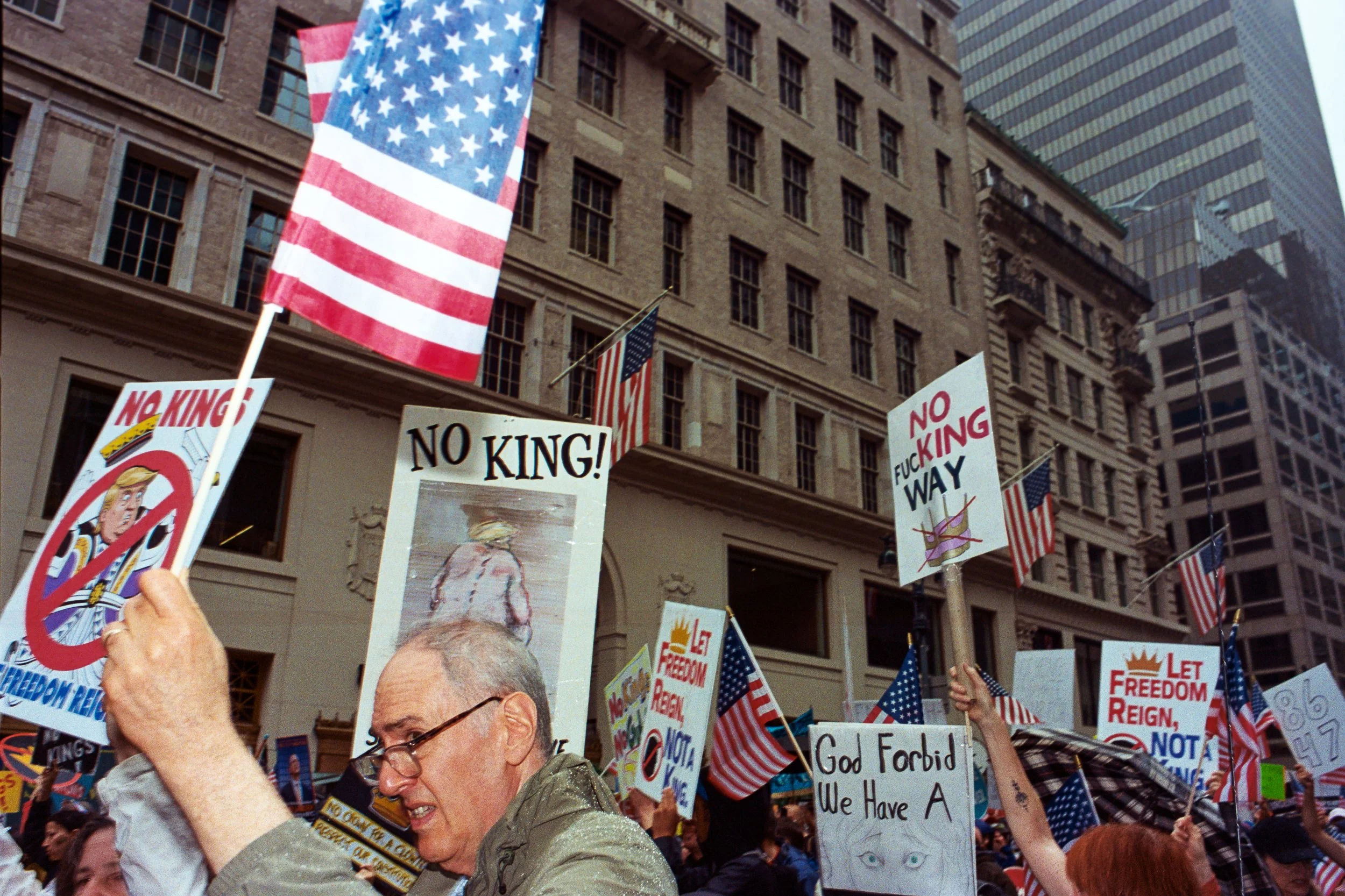 A crowd of protesters holding signs and American flags during a demonstration in an urban area with tall buildings.