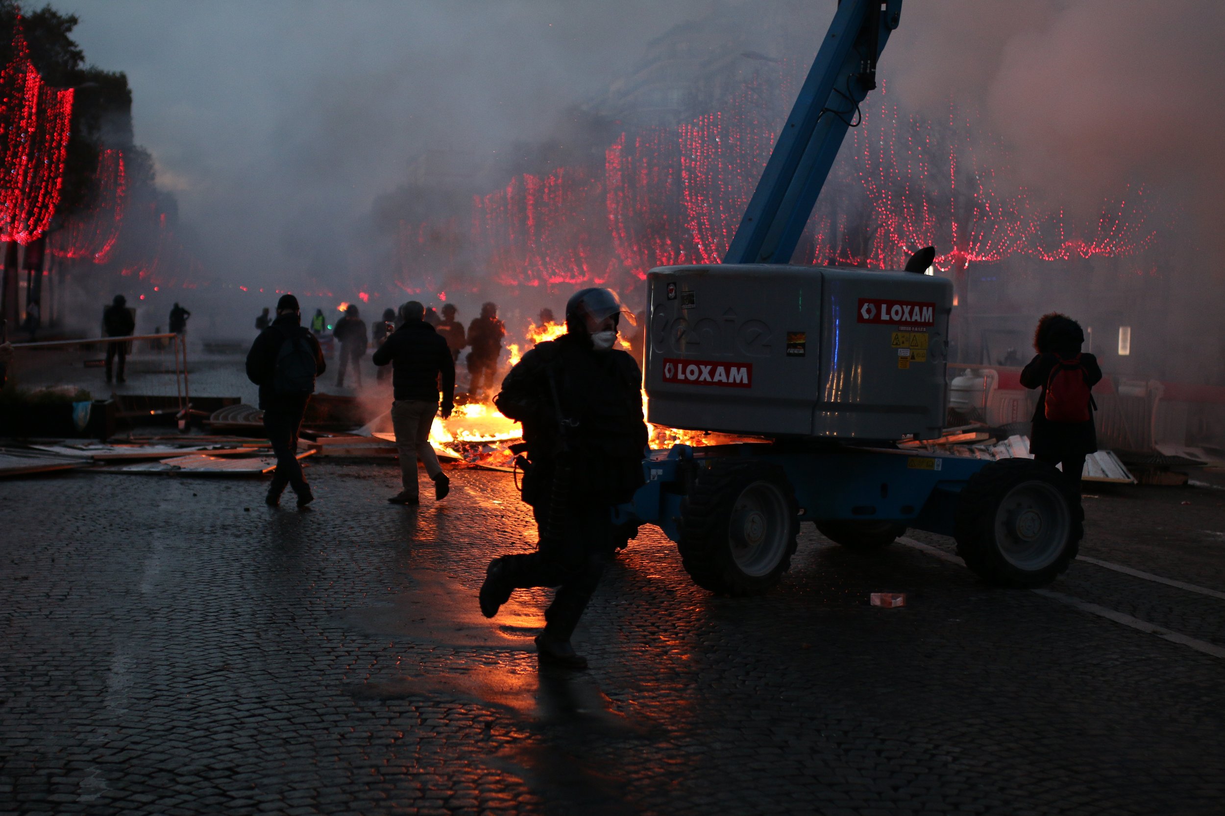People evacuating as a fire burns in a building, creating smoke and flames, with emergency personnel and equipment present.
