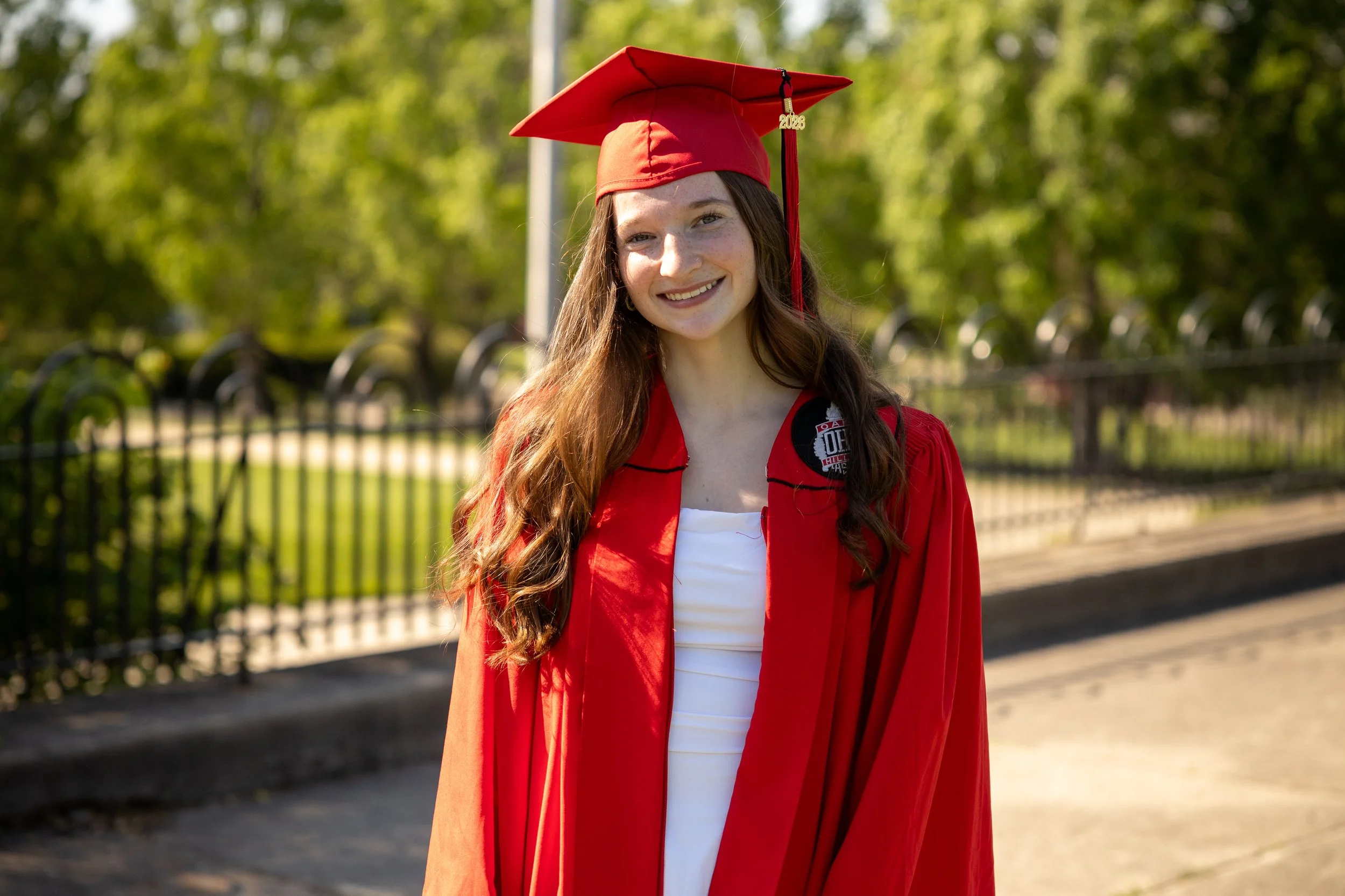 A young woman with long brown hair wearing a red graduation cap and gown, smiling outdoors with green trees and a black fence in the background.