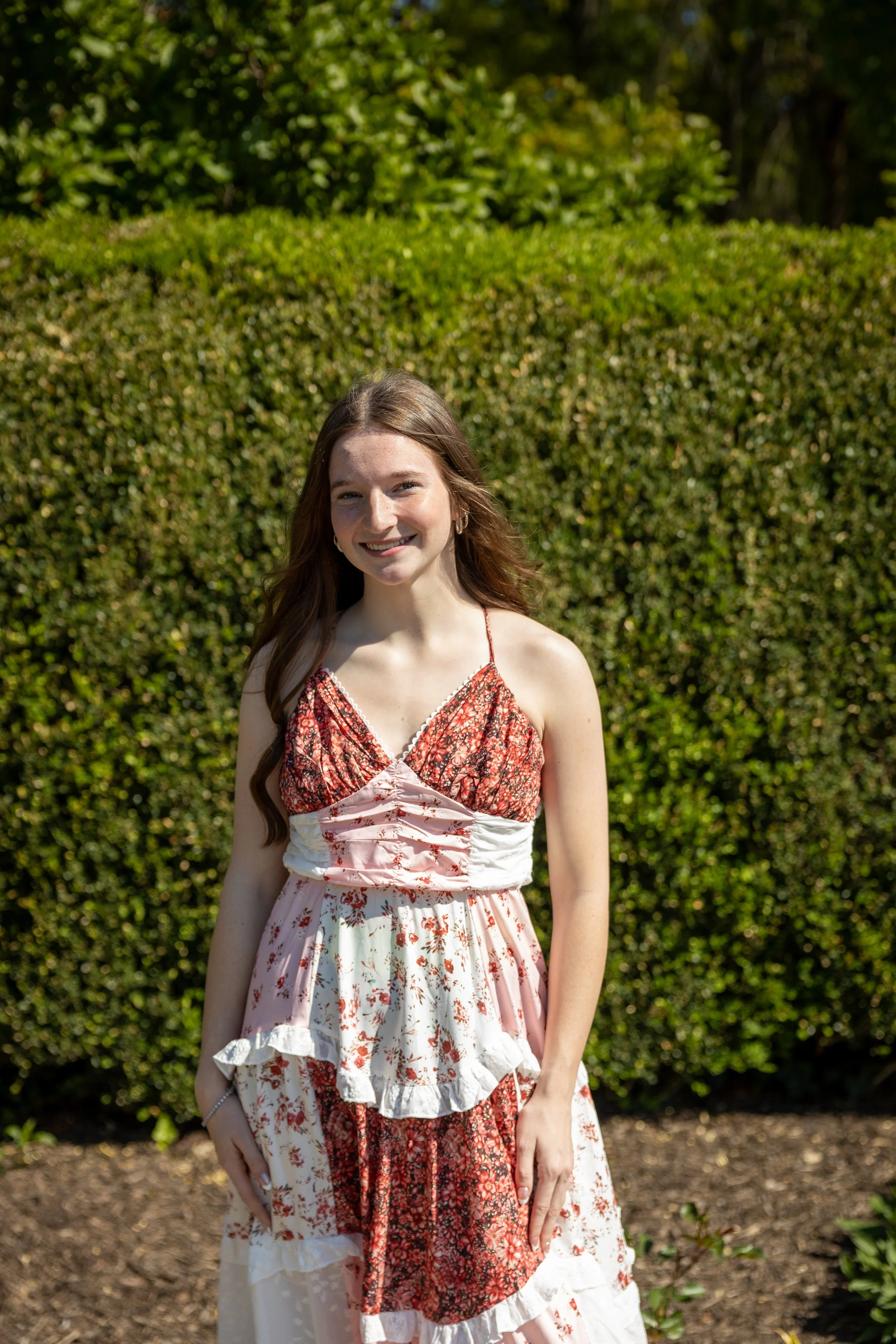 A young woman with long brown hair standing outdoors in front of a green bush, wearing a red and white floral dress, smiling at the camera on a sunny day.