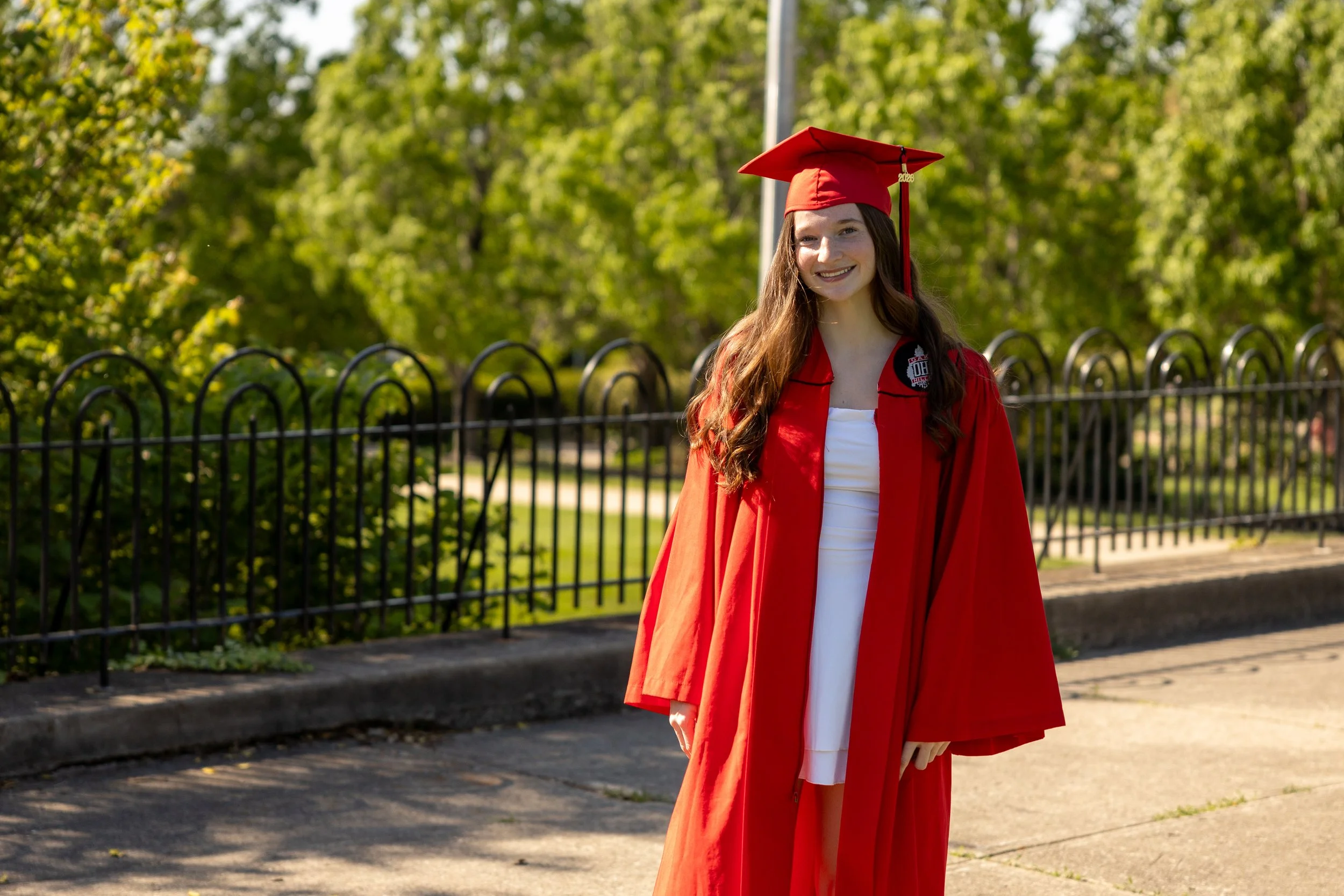 A young woman wearing a red graduation gown and cap with a tassel, standing outdoors in front of trees and a black metal fence, smiling.