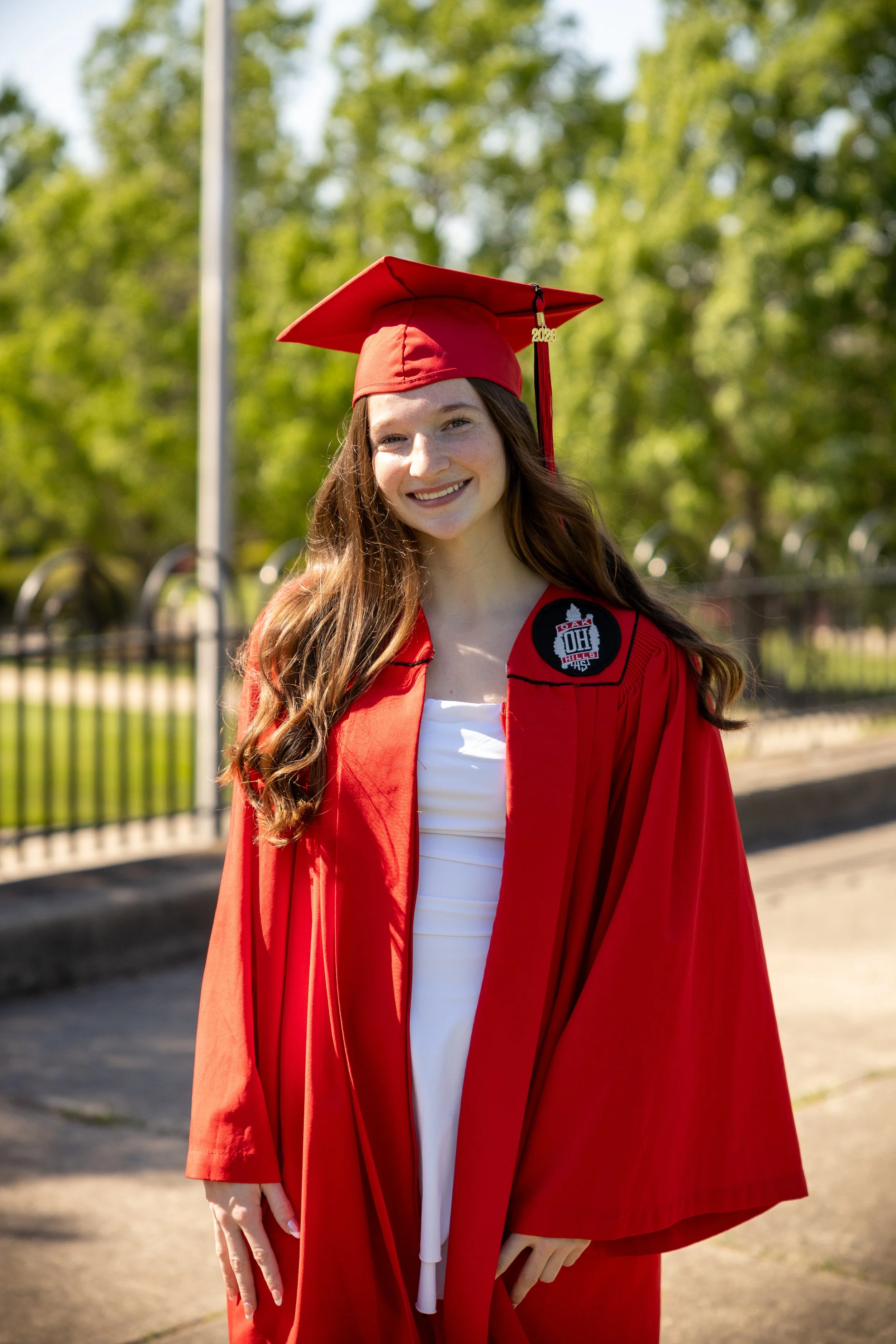 A young woman in a red graduation gown and cap with a tassel that says '2023', smiling outdoors against a background of trees and a fence.