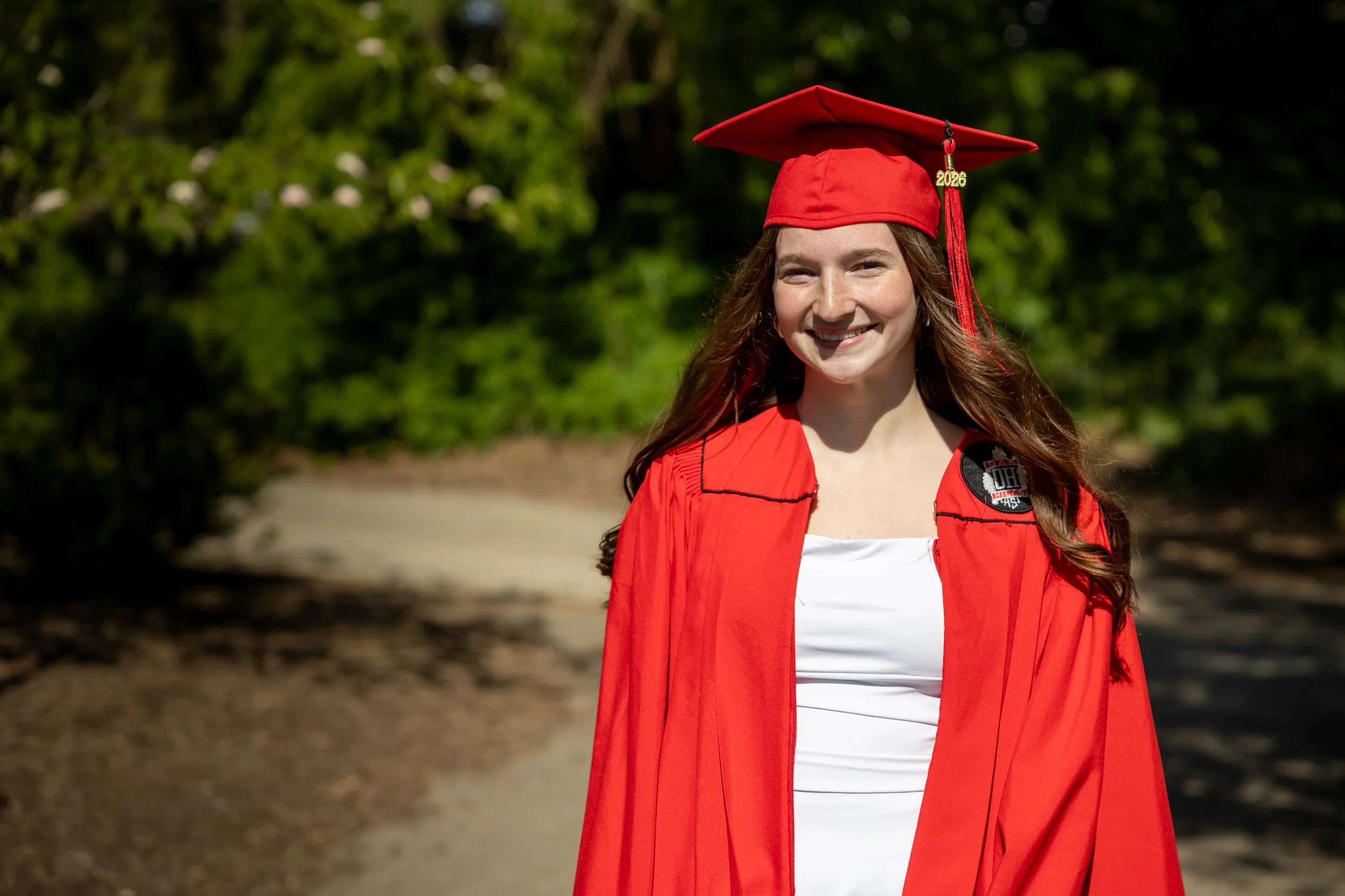 Young woman in red graduation cap and gown standing outdoors in front of a green leafy background, smiling.