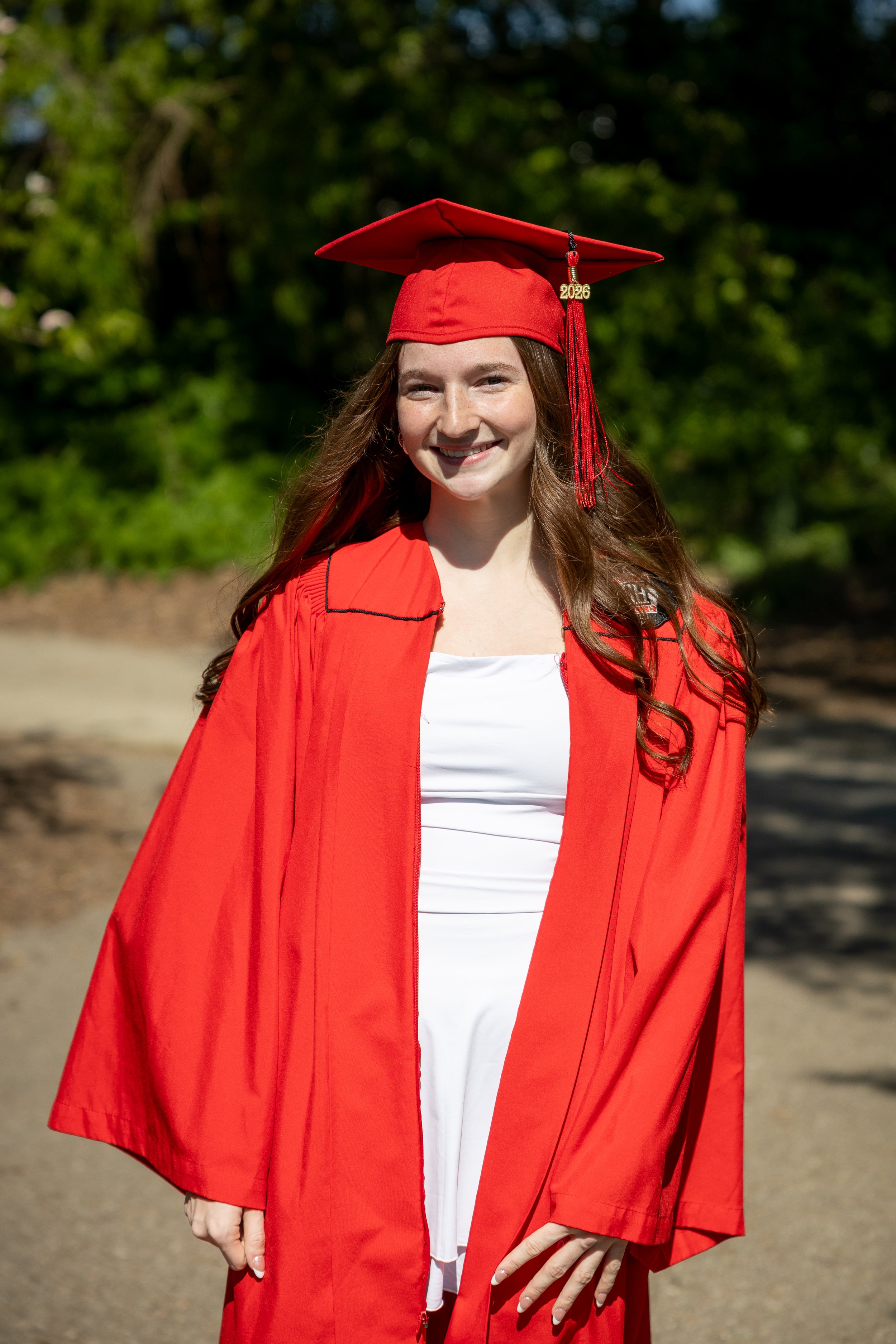 A young woman with long red hair smiling, wearing a red graduation cap and gown with a tassel marked 2026, in an outdoor setting with green trees in the background.
