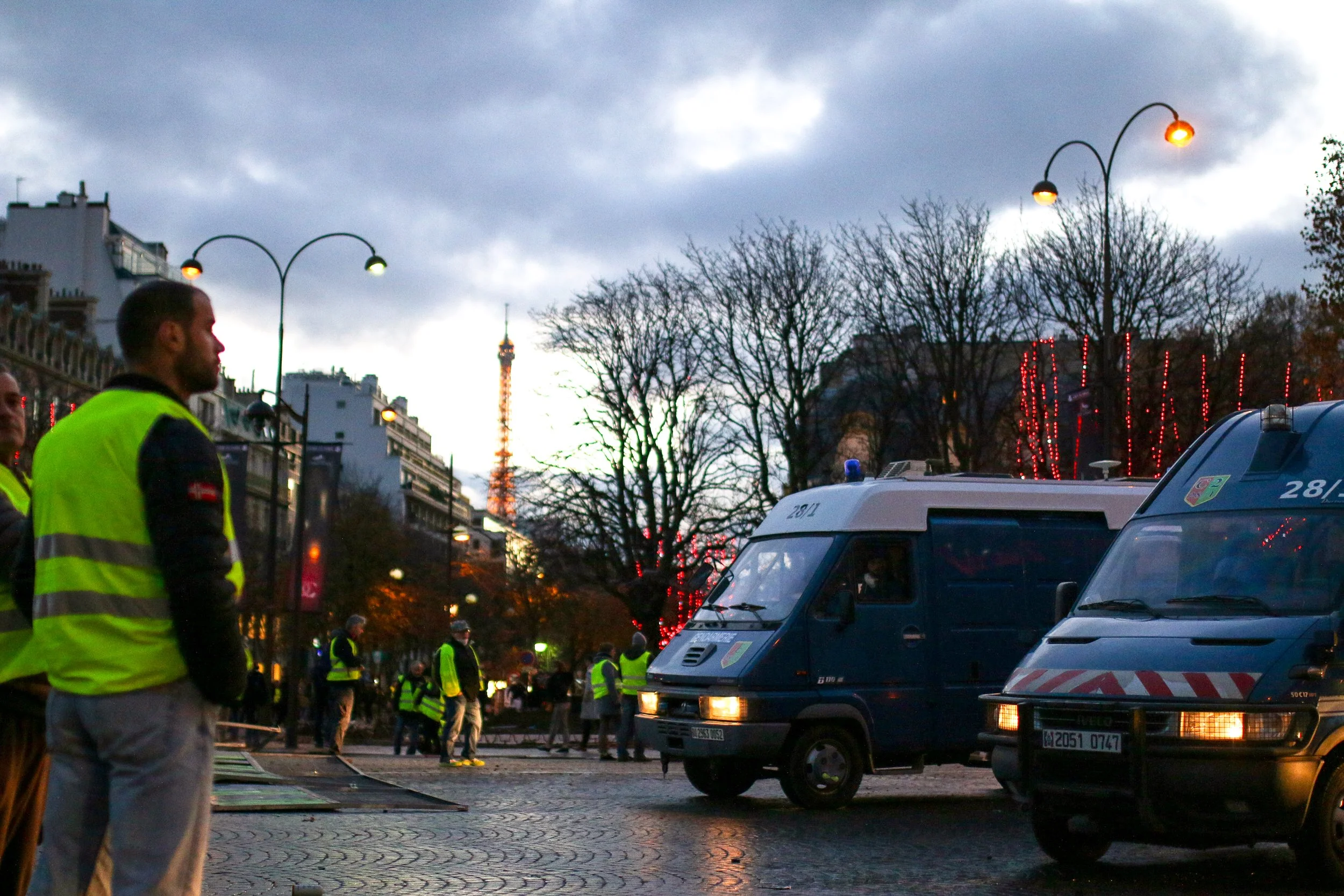 Paris city street scene during dusk with police and emergency vehicles, officers in yellow vests, bare trees, and the Eiffel Tower in the background.