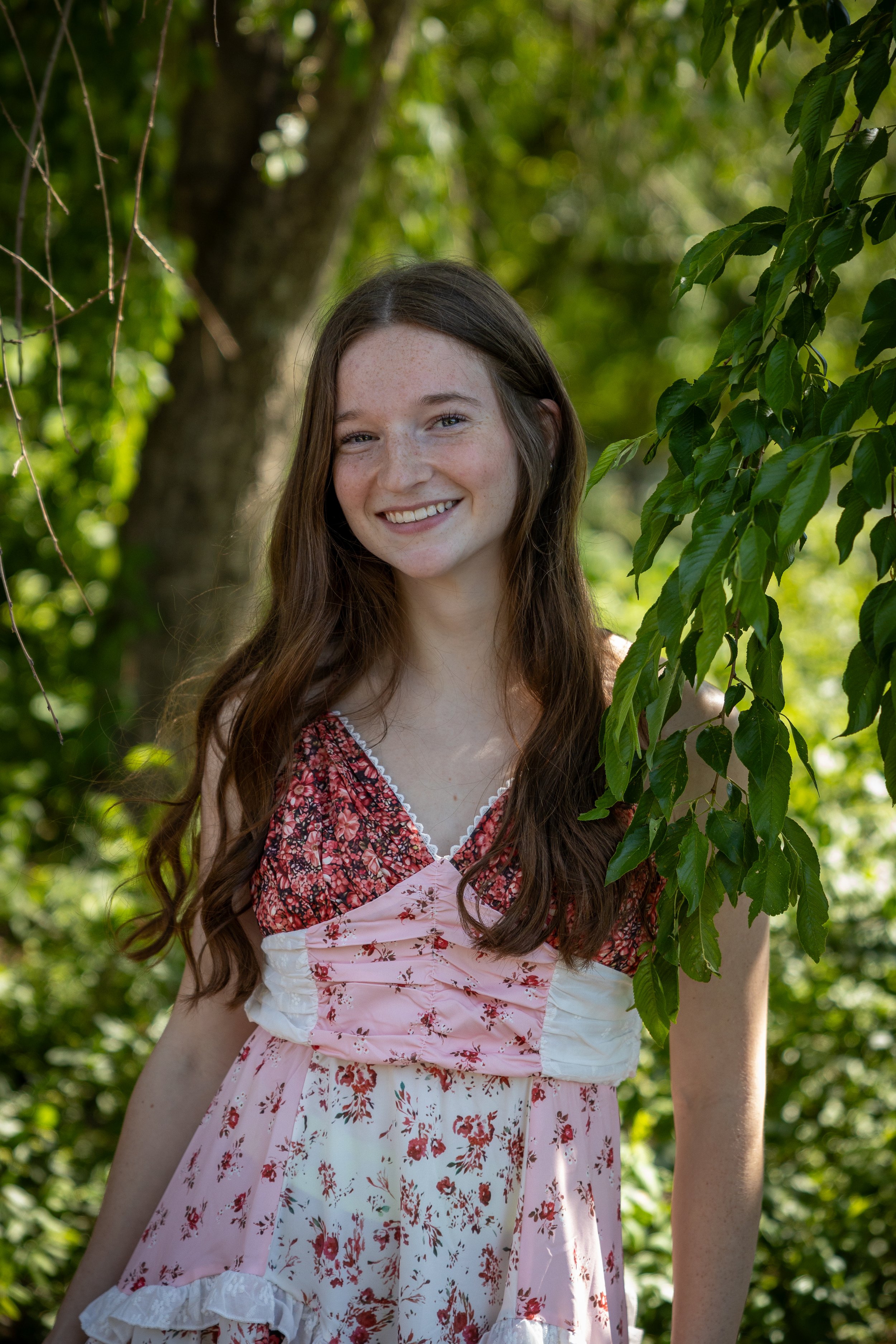 A young woman with long brown hair, smiling, standing outdoors among green trees, wearing a pink floral dress with ruffles.