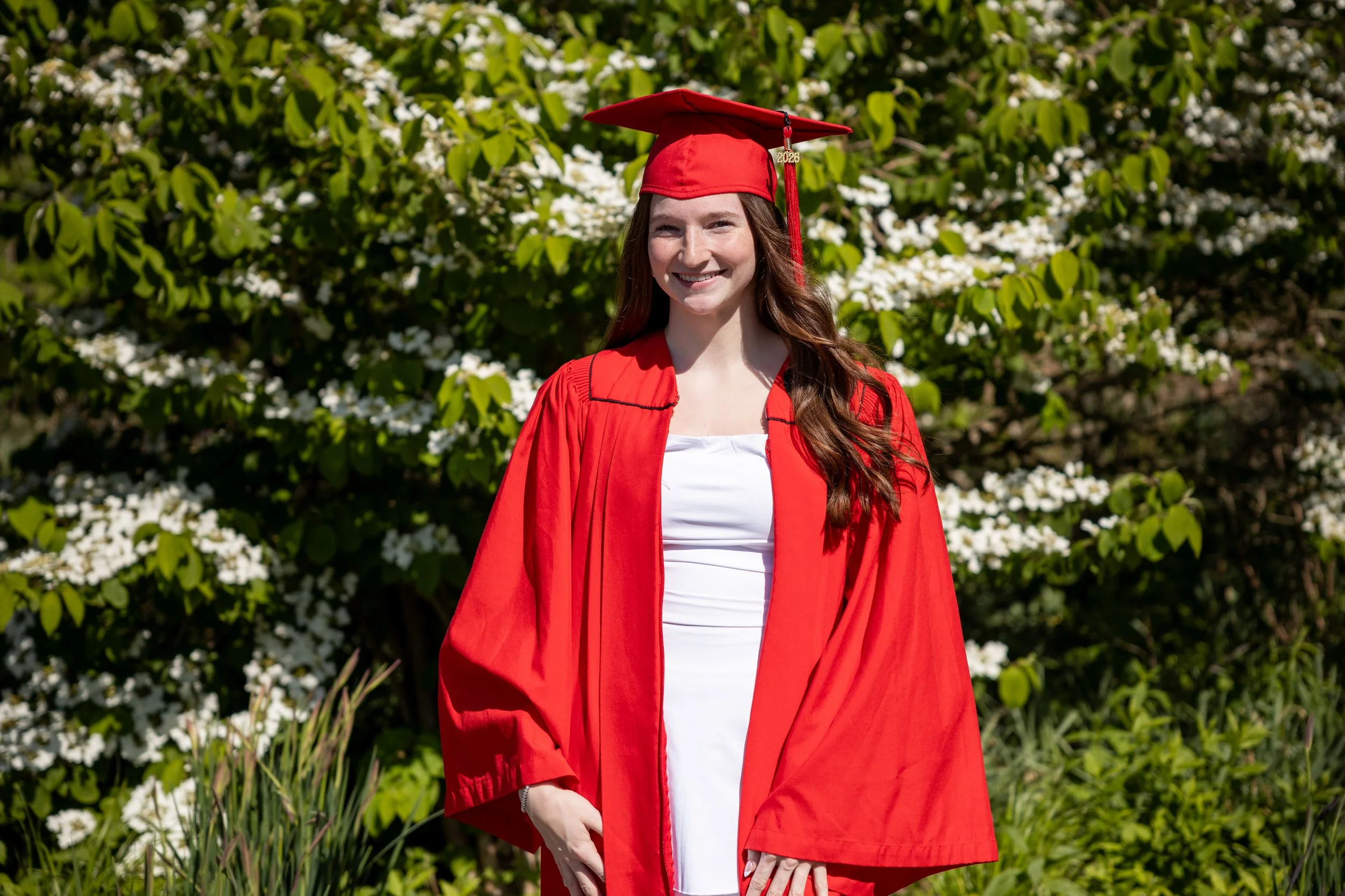 A young woman in a red graduation gown and cap, with a tassel marked 2026, smiling outdoors in front of green foliage and white flowers.
