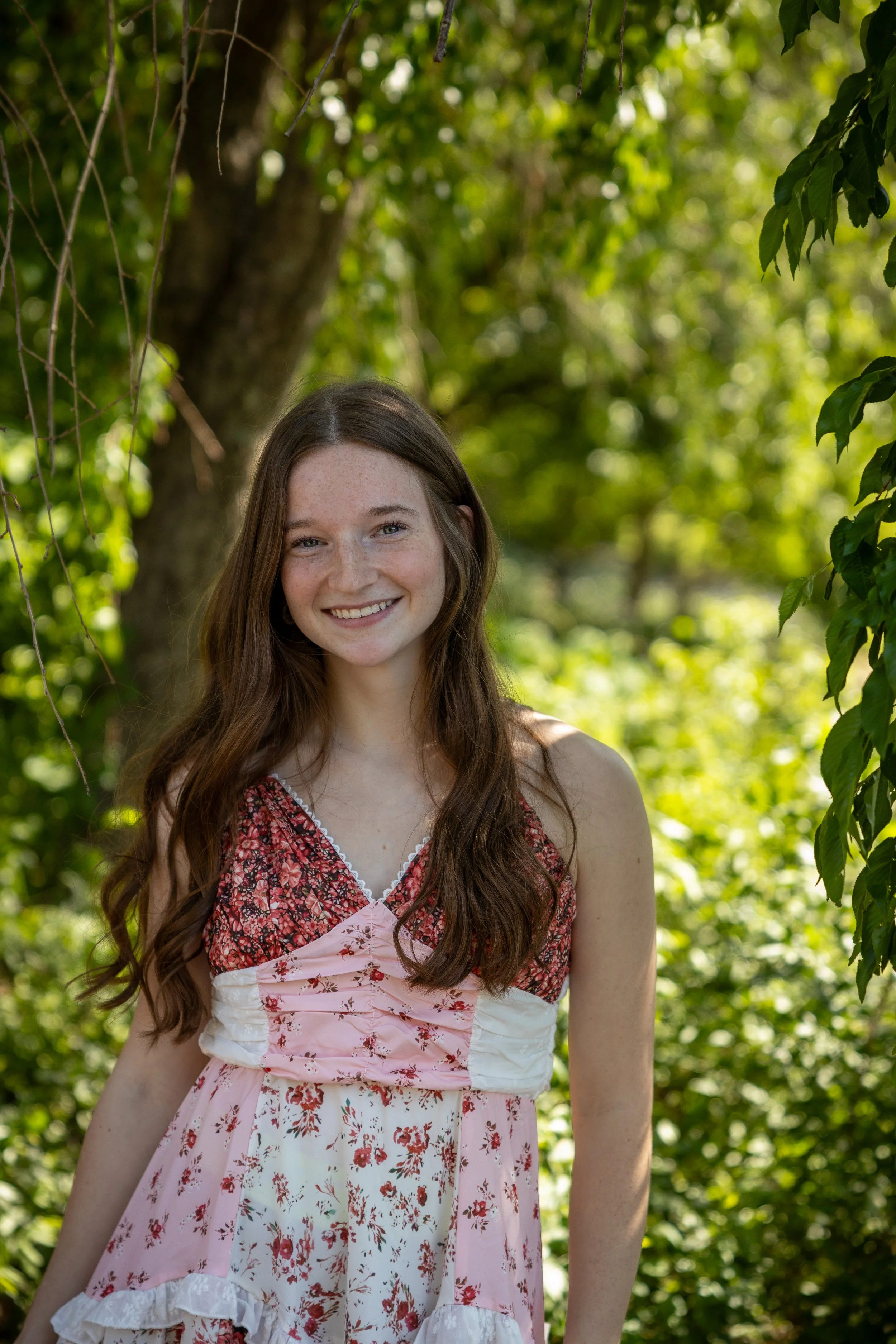 A young woman with long brown hair and freckles smiling outdoors, wearing a pink and red floral dress, surrounded by green trees and foliage in bright sunlight.