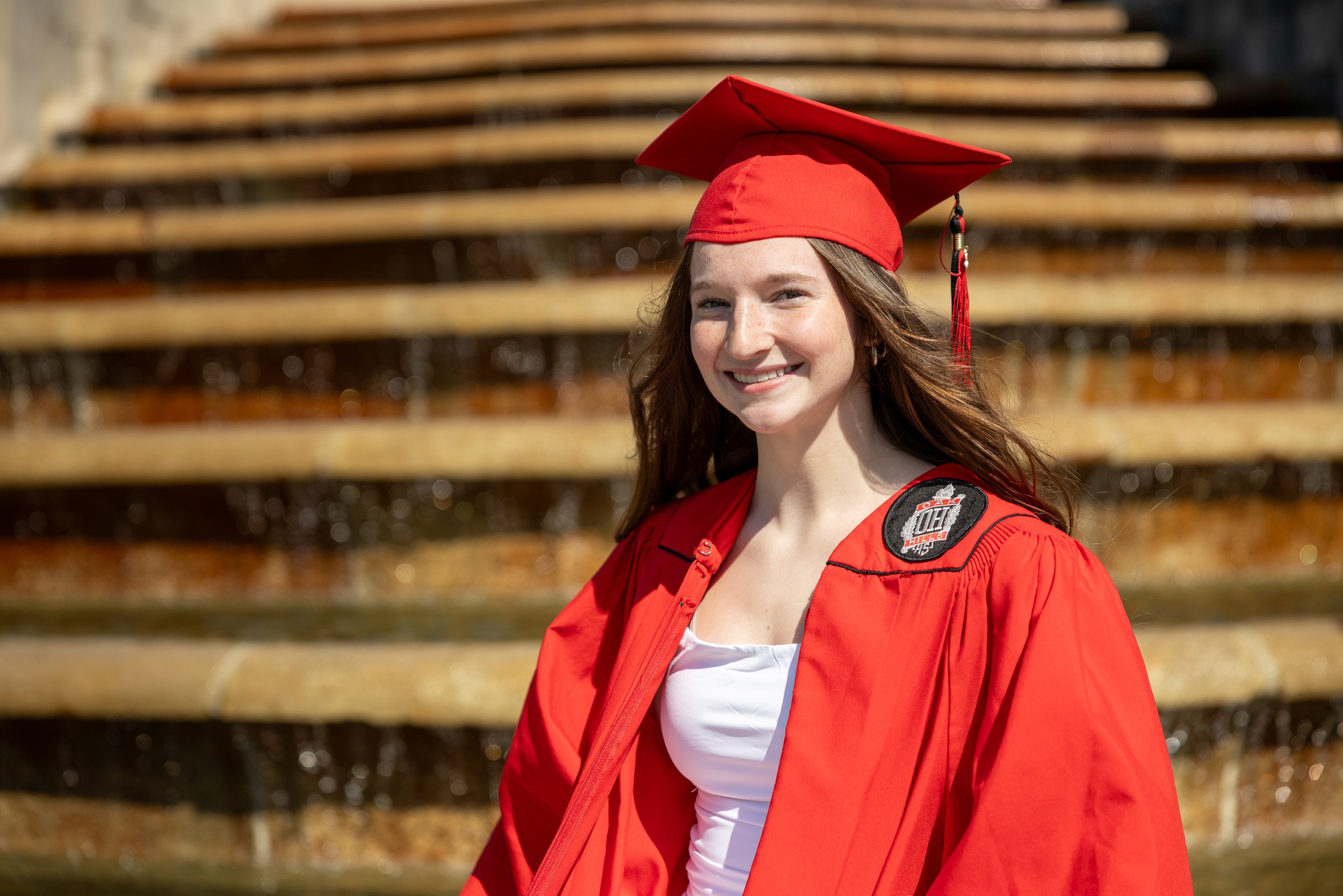 A young woman in a red graduation cap and gown smiling outdoors, with steps in the background.