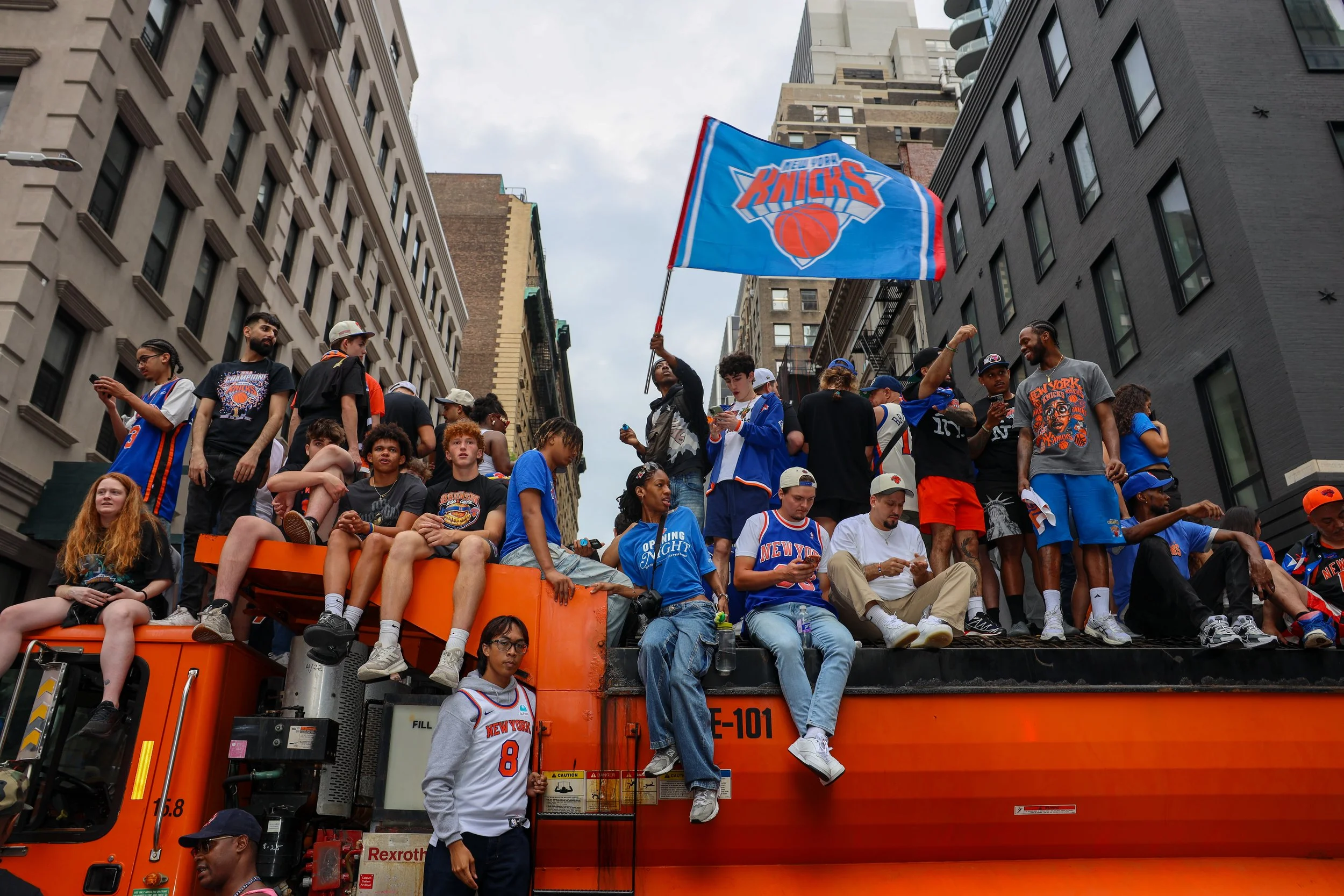 A parade float carrying New York Knicks basketball fans, with some sitting and some standing, surrounded by tall buildings in an urban setting. One person is waving a Knicks flag, and many are wearing Knicks jerseys and apparel.
