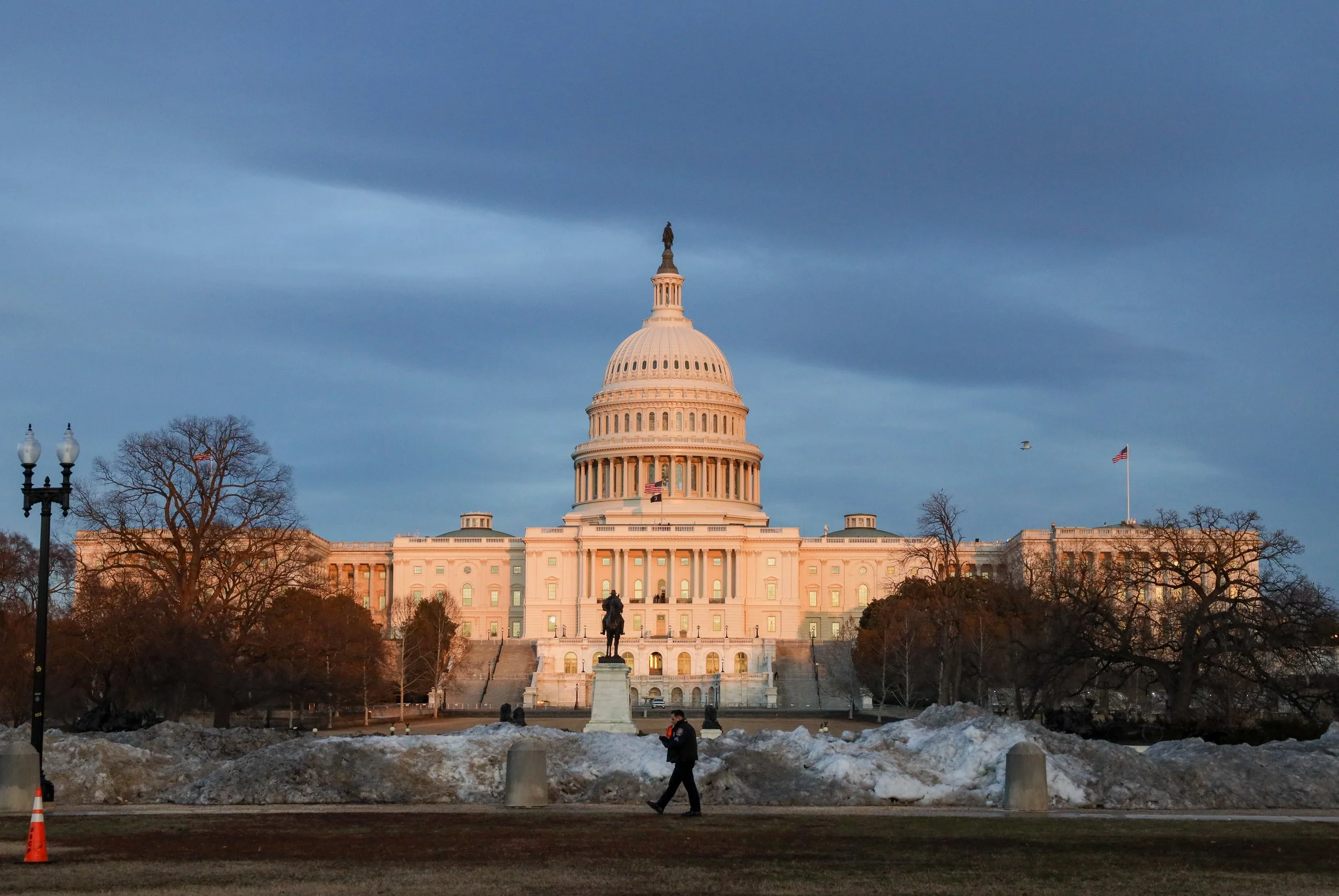 The United States Capitol building in Washington, D.C., during late afternoon or early evening. The sky is cloudy with a slight glow from the setting sun. There are a few trees in the foreground, some covered with snow, and a person walking nearby. A