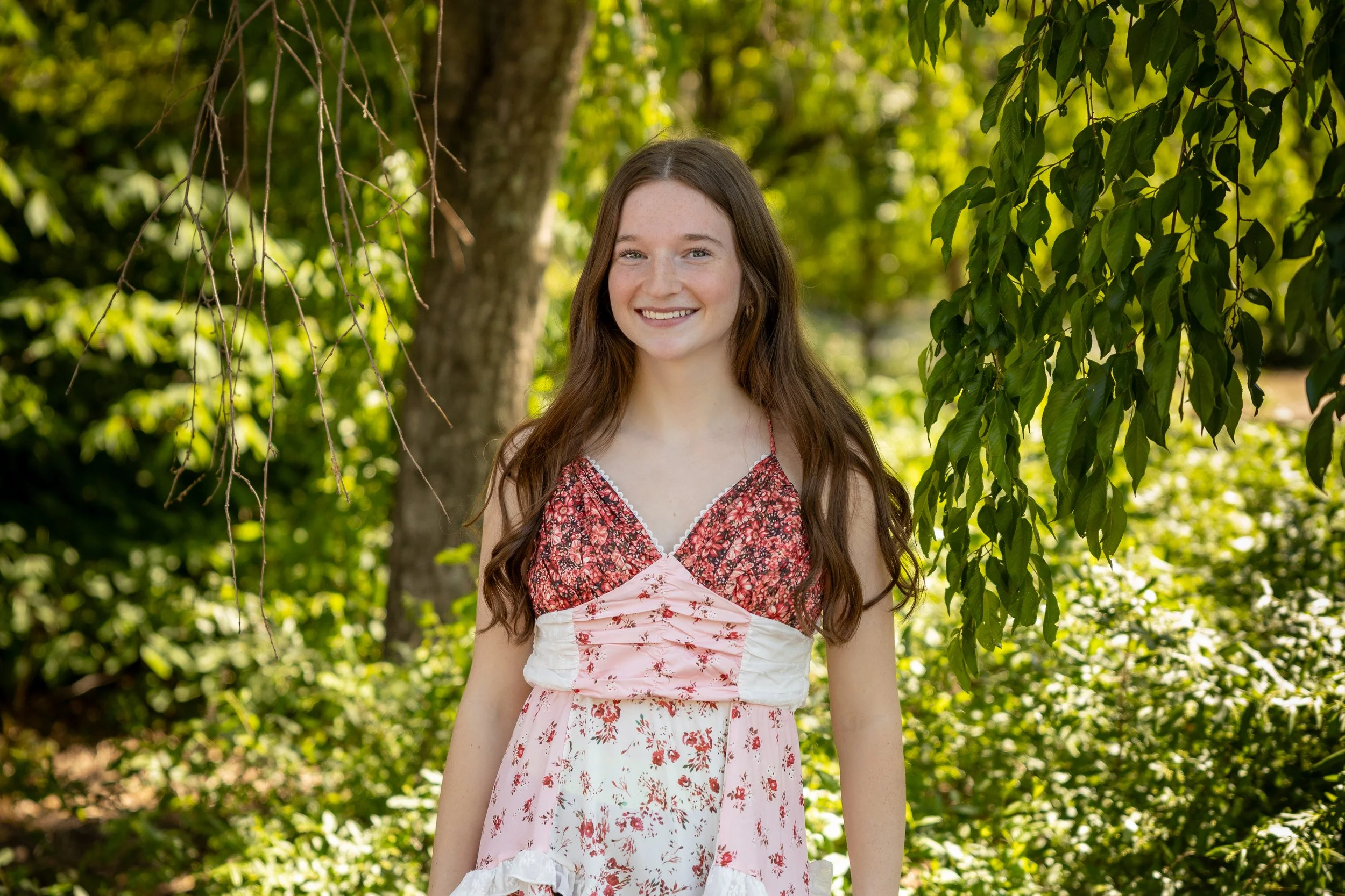 A young woman with long brown hair smiling outdoors, wearing a pink and red patterned dress, with green trees and leaves in the background.