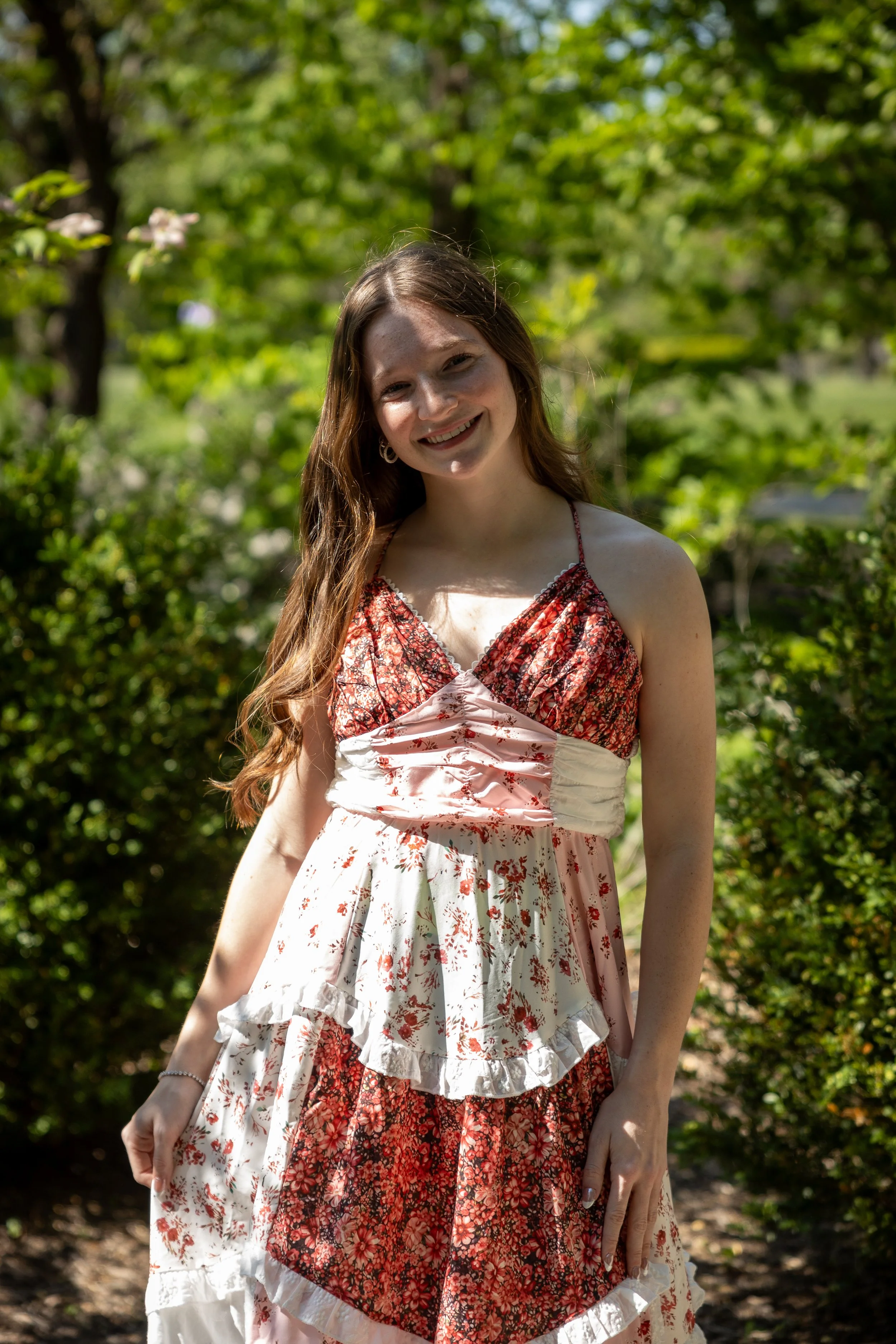 A young woman with long brown hair, smiling, wearing a floral dress, standing outdoors on a sunny day with green trees and bushes in the background.
