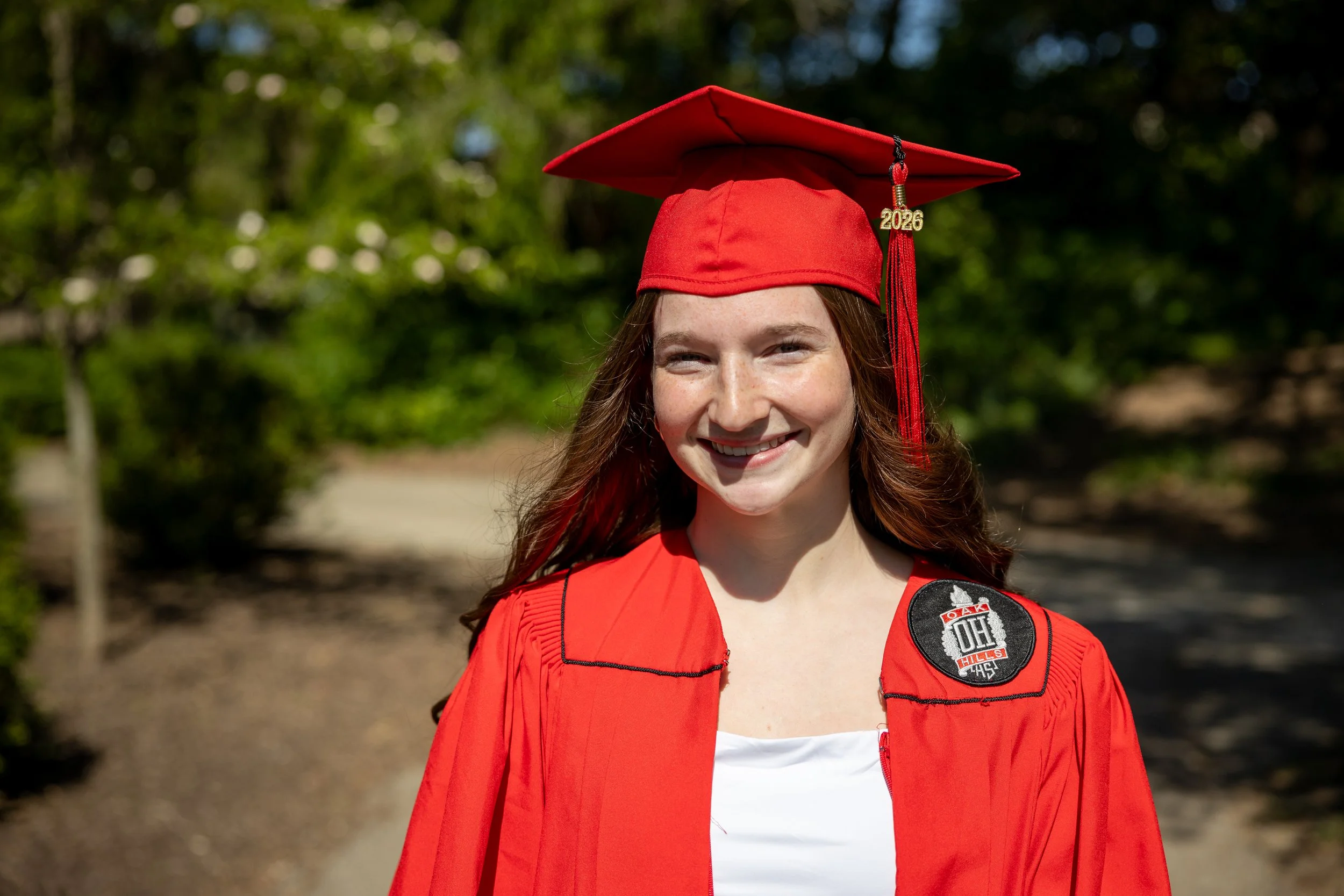 Young woman with red hair smiling outdoors, wearing a red graduation cap with a gold '2026' tassel and a red jacket with patches.