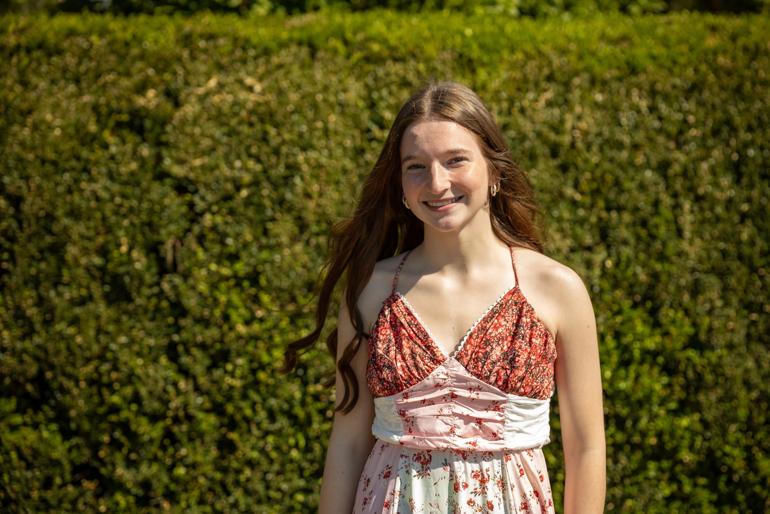 A young woman with long red hair smiling outdoors in front of a green hedge, wearing a red patterned sleeveless dress.