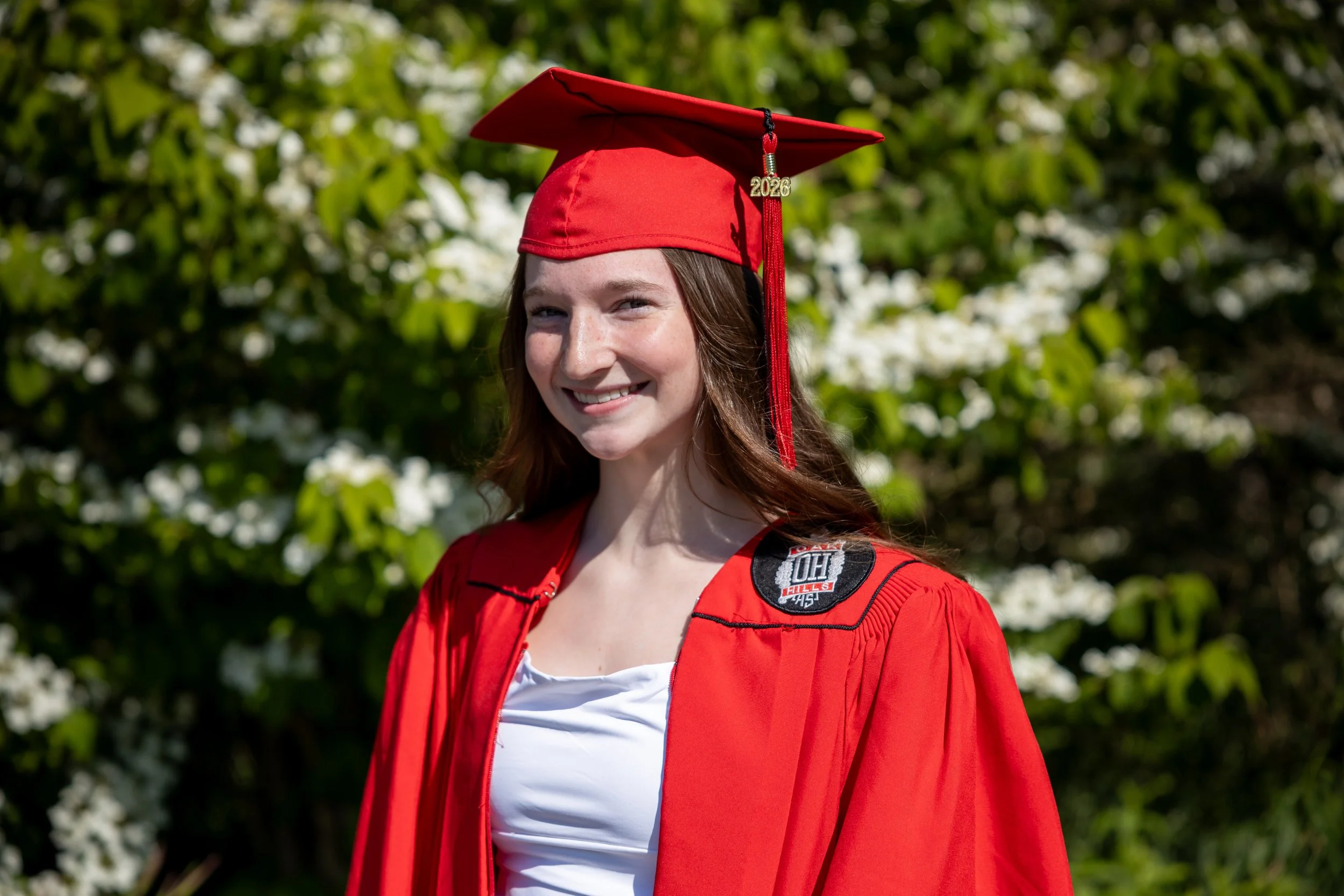 A young woman in a red graduation gown and cap decorated with a '2026' charm, smiling outdoors with green foliage in the background.