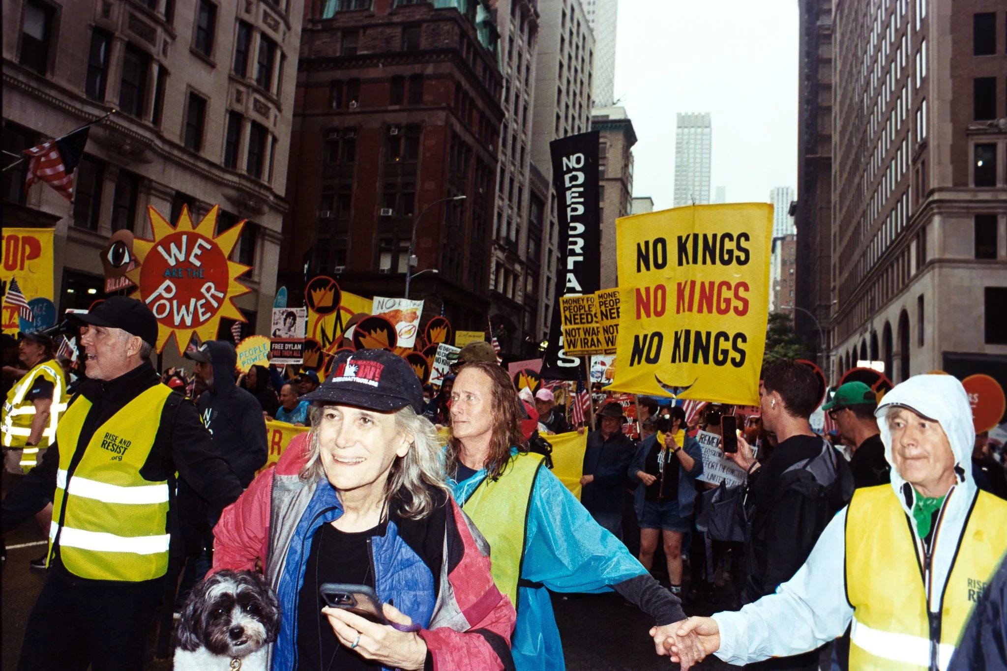 A group of protesters marching in a city street surrounded by tall buildings, holding yellow signs with messages like "No Kings" and "We Are The Power," wearing yellow vests and rain gear, with some protesters holding flags and a woman with a dog in 