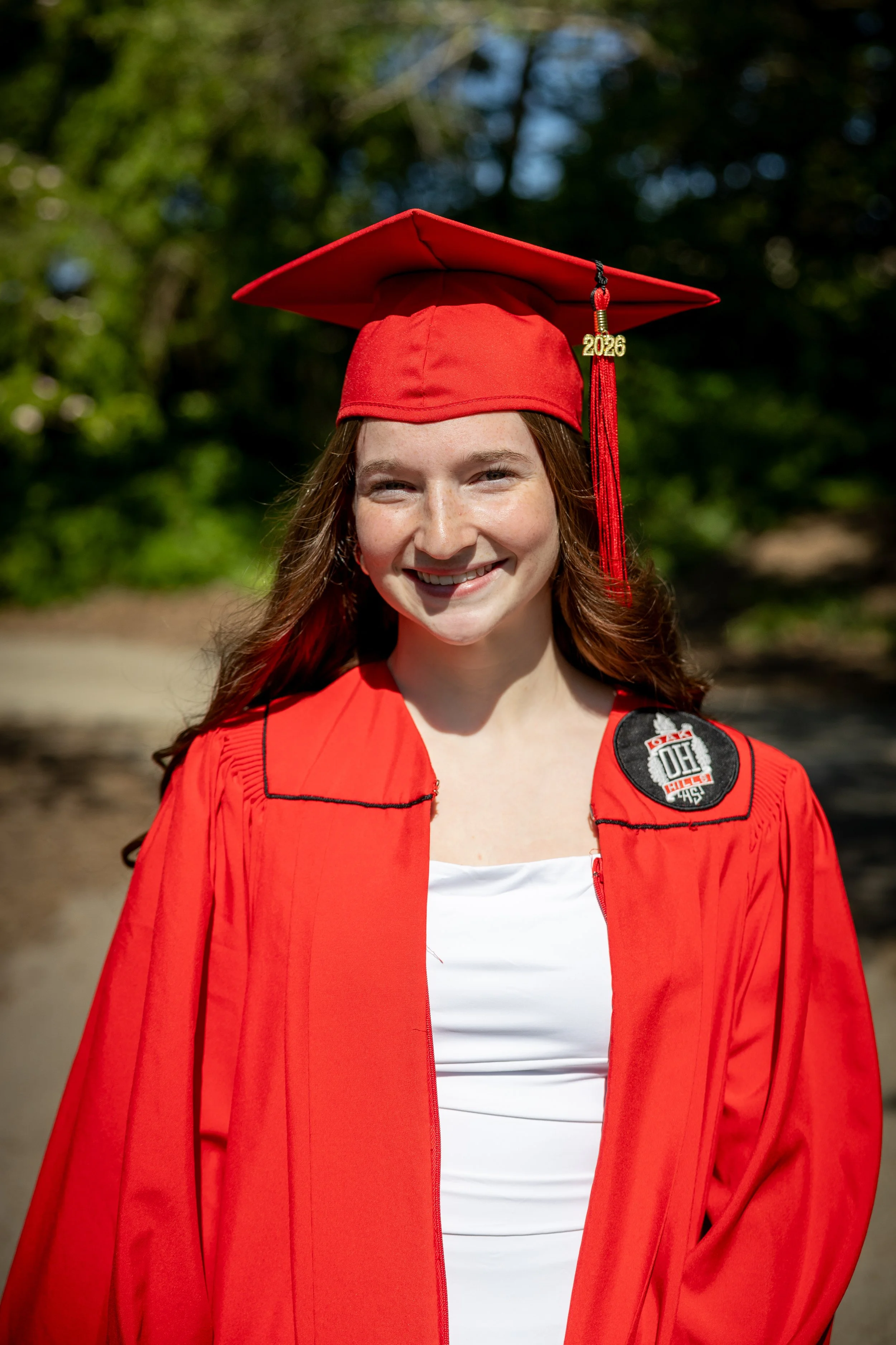 A young woman with long brown hair smiling outdoors, wearing a red graduation cap with '2026' charm and a red graduation gown over a white top, with a background of blurred green trees and sunlight.