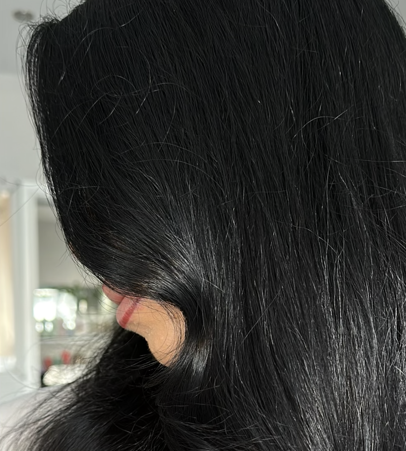 Close-up of a person's head showing long, straight, black hair, with a small section tucked behind the ear, in a bright indoor setting.