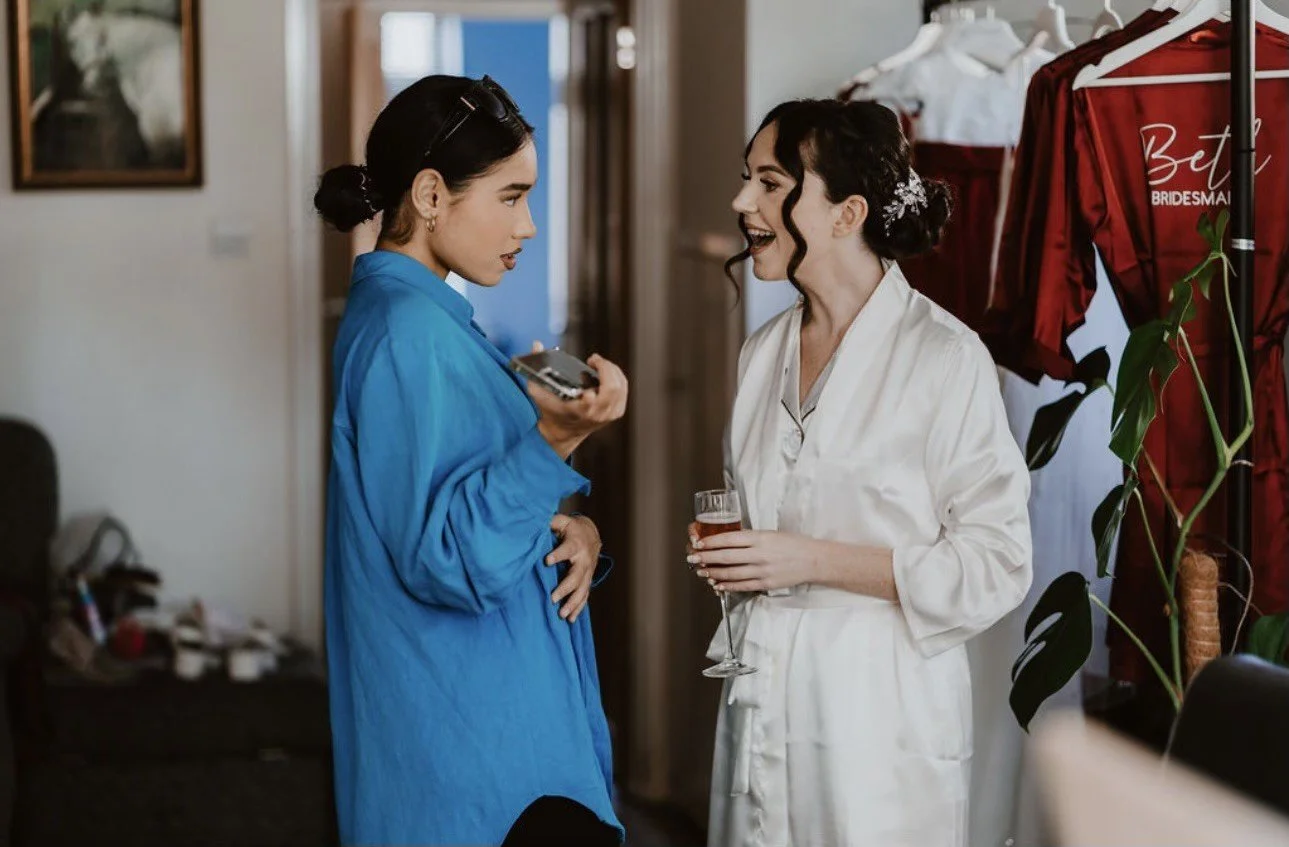 Two women are having a conversation in a dressing room area. One is in a blue robe and the other in a white robe, holding a glass of champagne. There are wedding dresses hanging in the background.