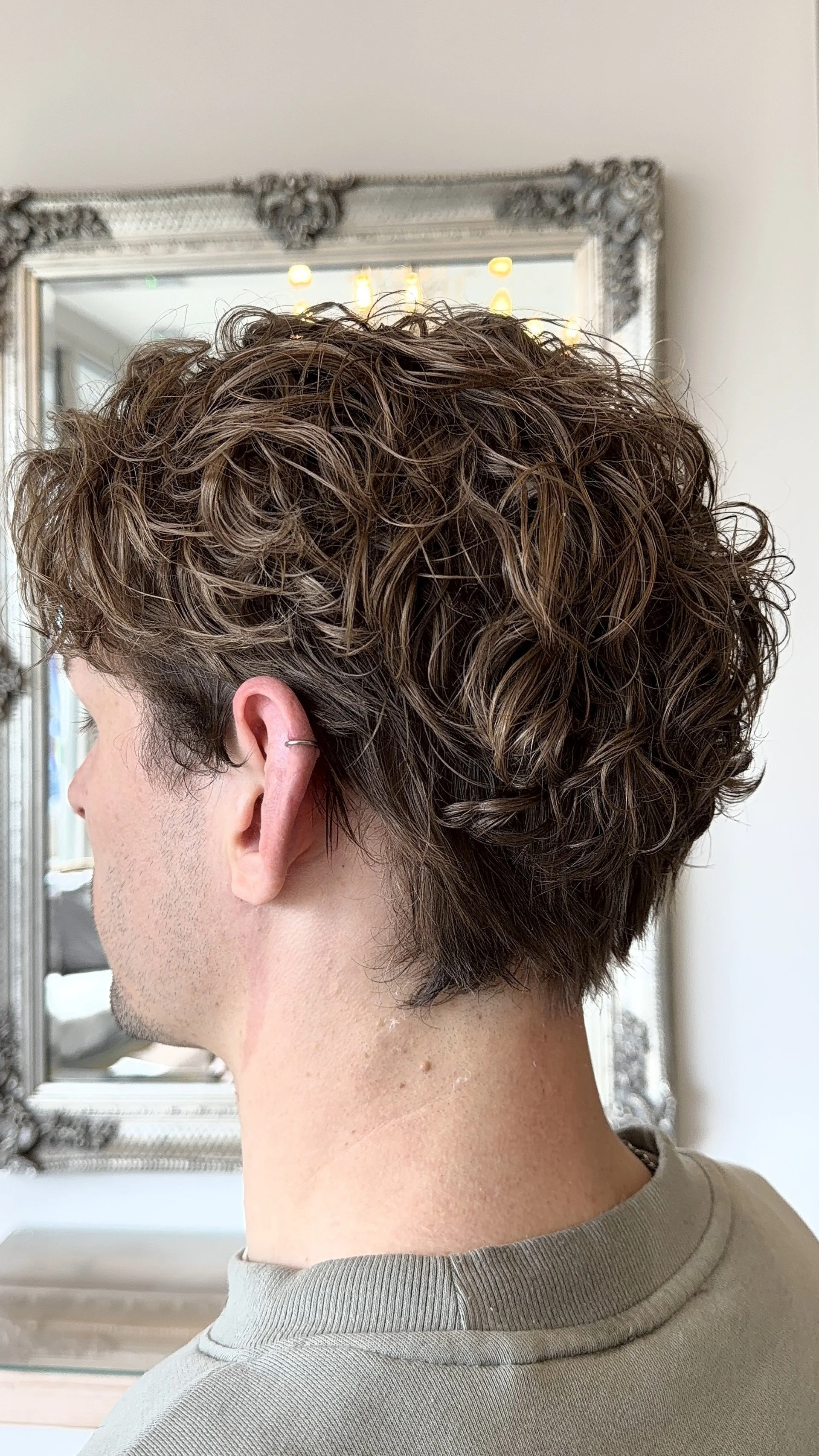 A person with short, curly brown hair viewed from the side, standing in front of a mirror.
