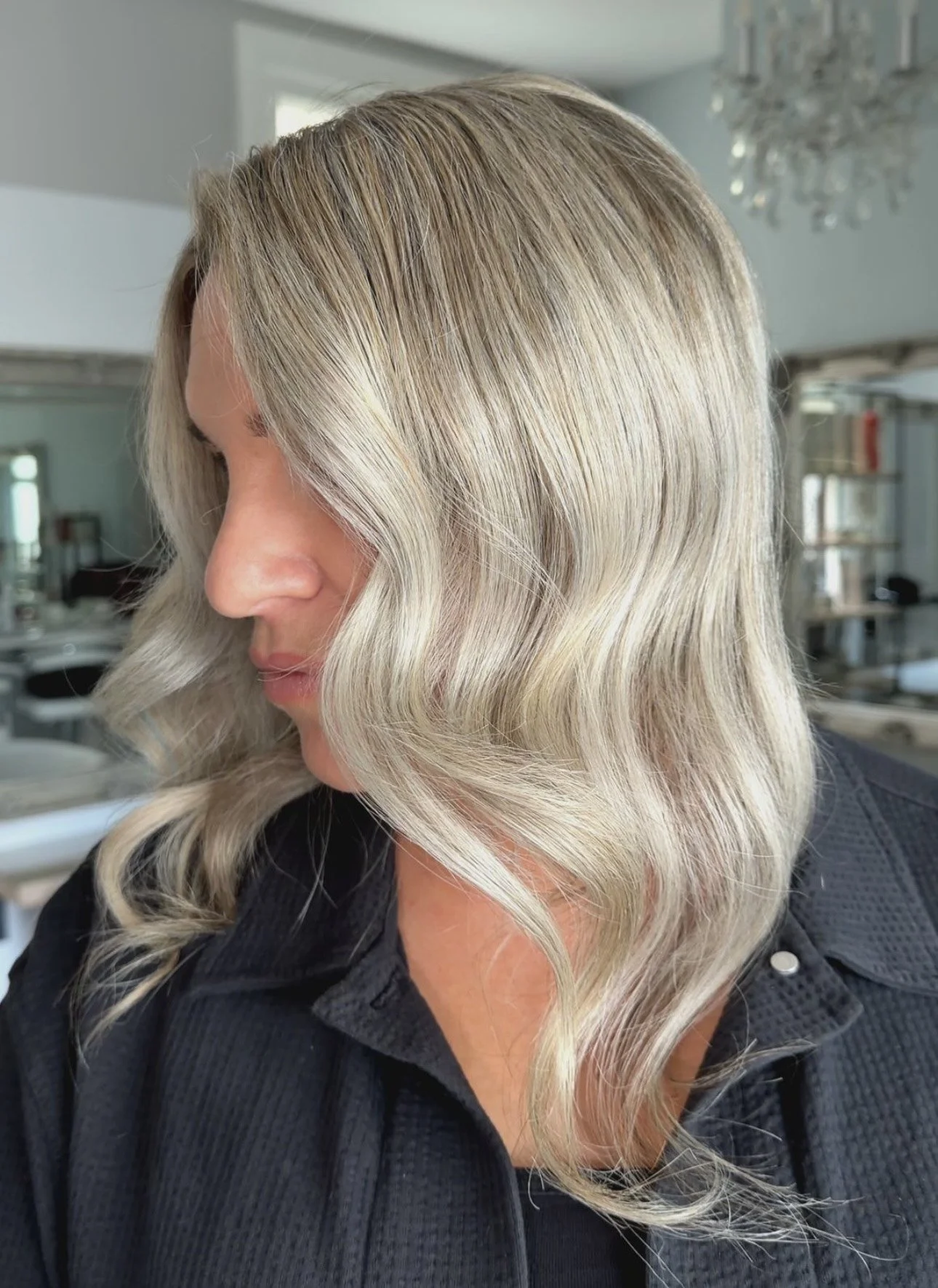Close-up of a woman with blonde, wavy hair styled in soft curls, wearing a black textured jacket, in a room with bright lighting and a chandelier in the background.