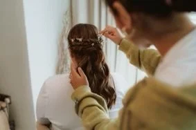 Back view of a woman with long, wavy, blonde hair wearing a brown sweater, standing indoors in front of a mirror.