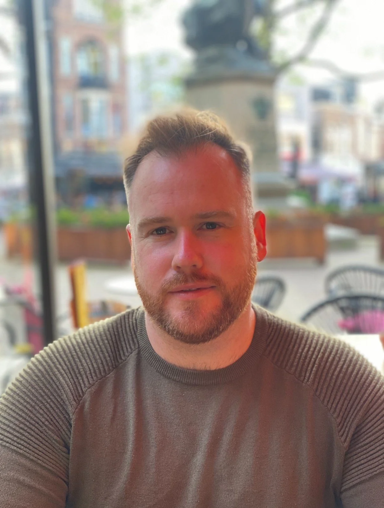 A man with short, light brown hair, a beard, and wearing a brown sweater, sitting inside a restaurant with a large window behind him. The background outside the window shows a city street with buildings, trees, and outdoor seating.