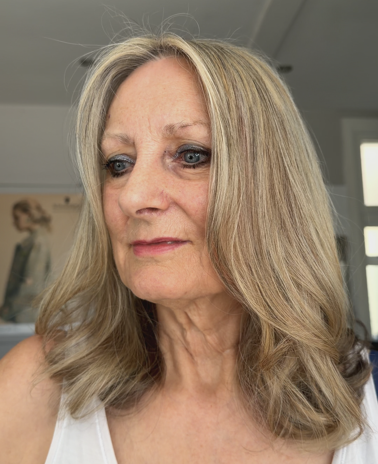 A woman with long blonde wavy hair is looking slightly to her right. She has blue eyes and is wearing makeup, including eyeliner and lipstick. She is dressed in a white top, and the background shows a room with a blurred poster or photo and a window.