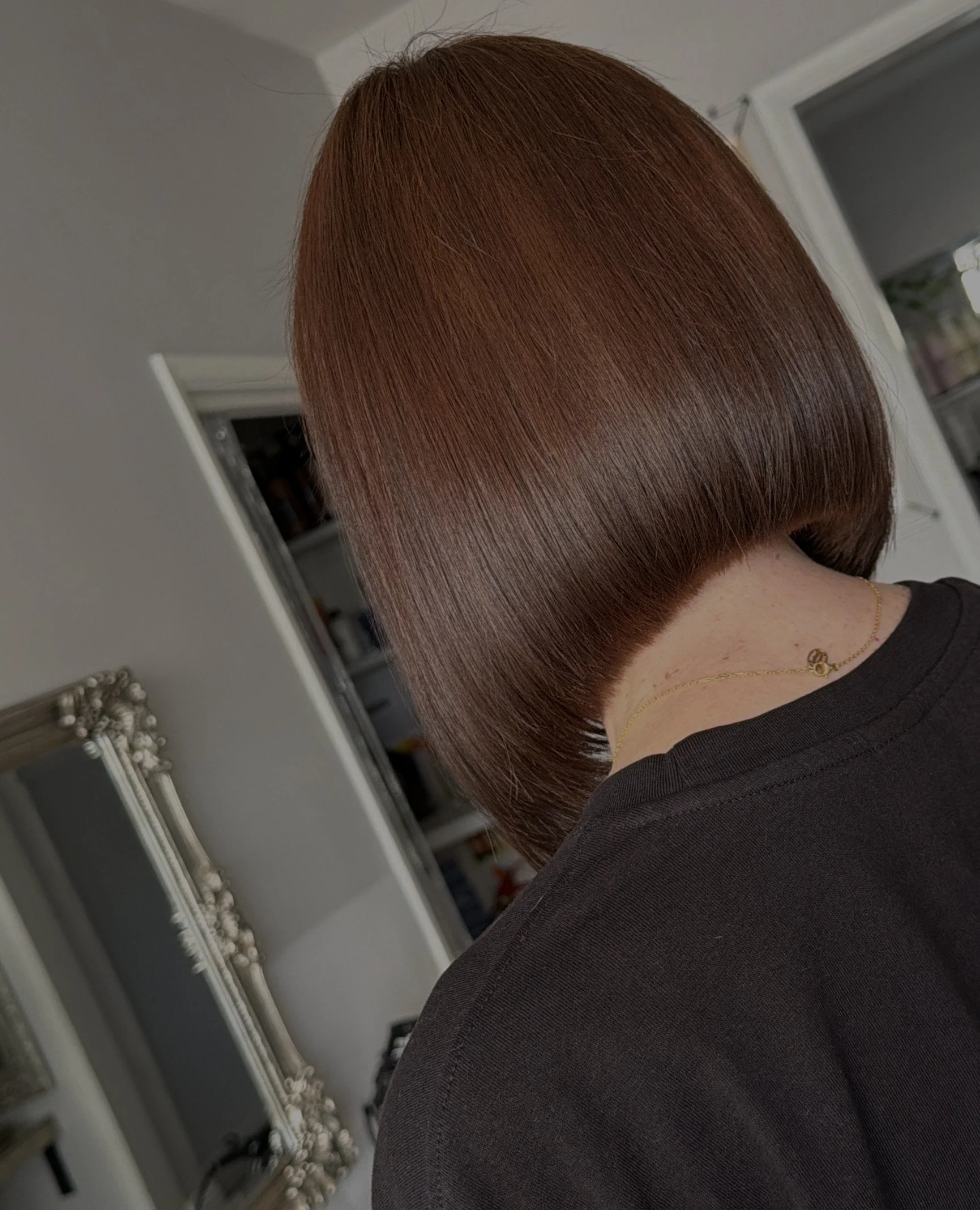 Close-up of a person's head with short, reddish-brown hair in a bob style, wearing a black top and a gold necklace, indoors near a window and shelves.