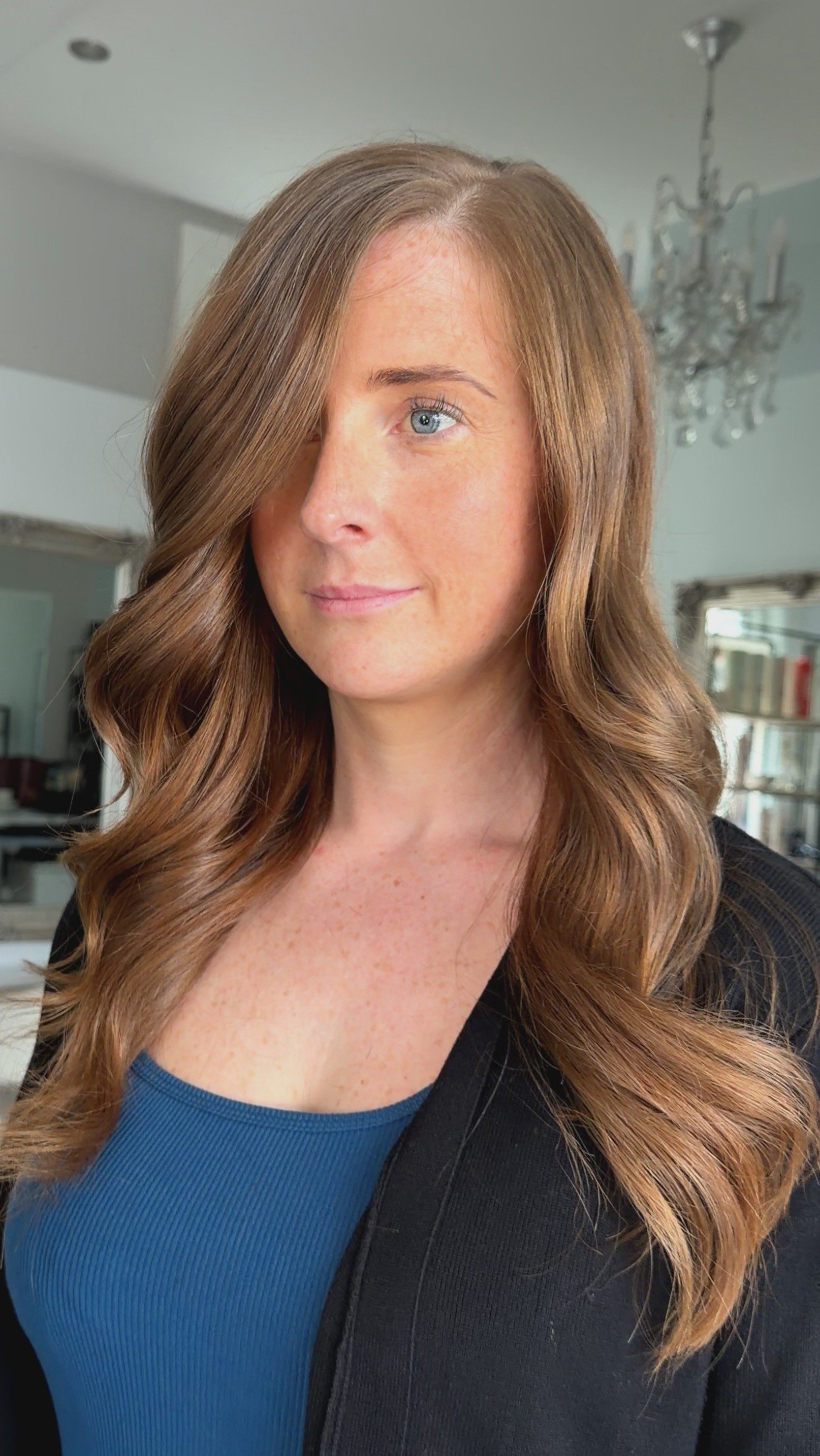 A woman with long, wavy, reddish-brown hair, wearing a black jacket over a blue top, standing in a room with a chandelier and bookshelf in the background.