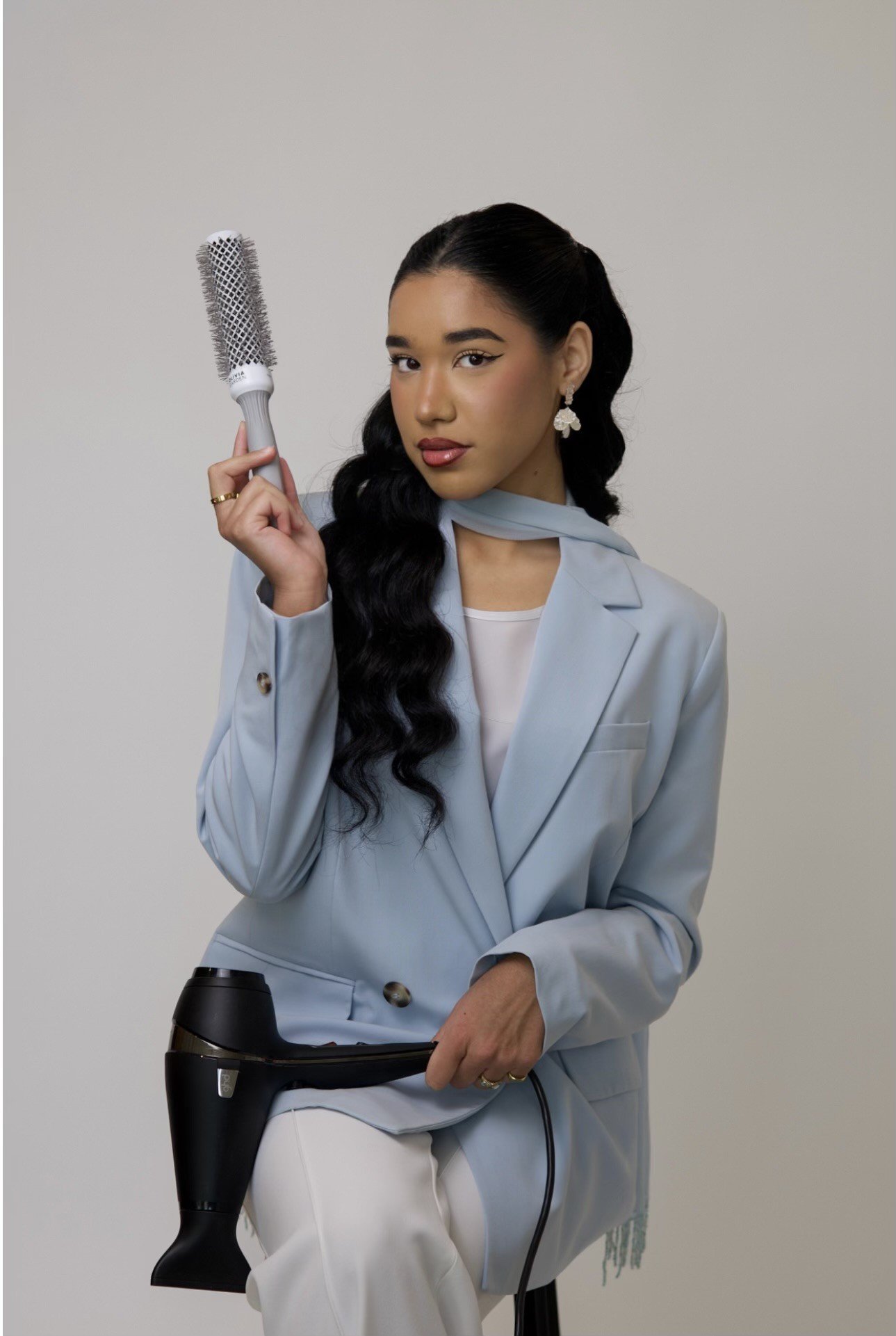 A woman with long dark wavy hair, wearing a light blue blazer and white pants, holding a hairbrush in one hand and a hairdryer in the other.