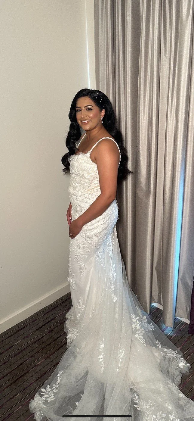 A woman in a white wedding dress standing next to beige curtains in a room with wooden flooring, smiling at the camera.
