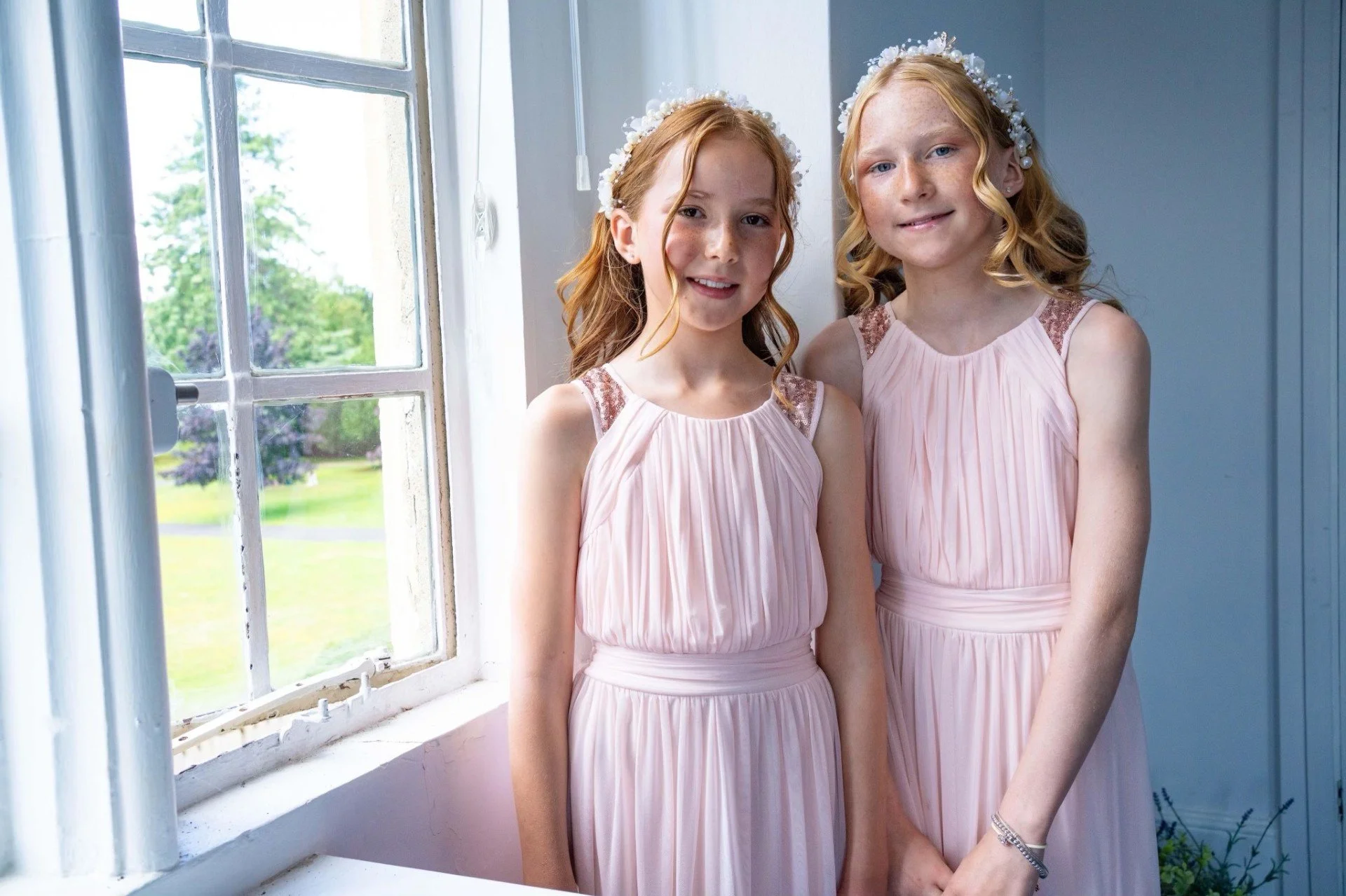 Two young girls with red hair wearing matching pink dresses and floral headbands standing indoors near a window.