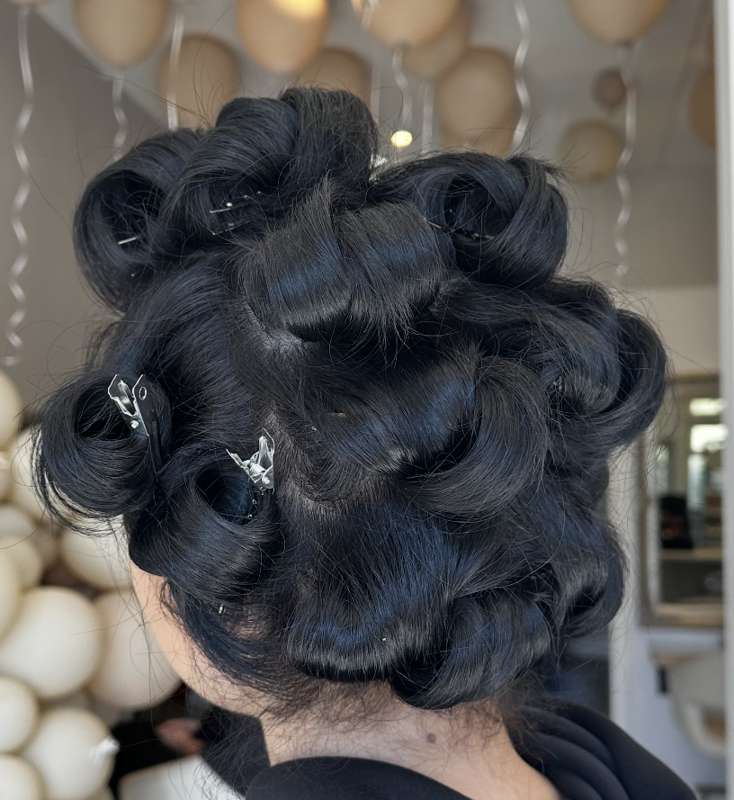 Back of a woman's head with hair set in large black curls, secured with silver clips, in a salon with balloon decorations in the background.