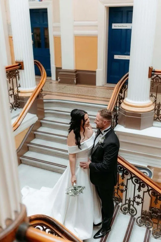 A bride and groom stand on a staircase inside a building, gazing into each other's eyes. The bride wears a white off-the-shoulder wedding gown and holds a bouquet, while the groom is dressed in a black suit with a tie. The staircase has ornate iron r