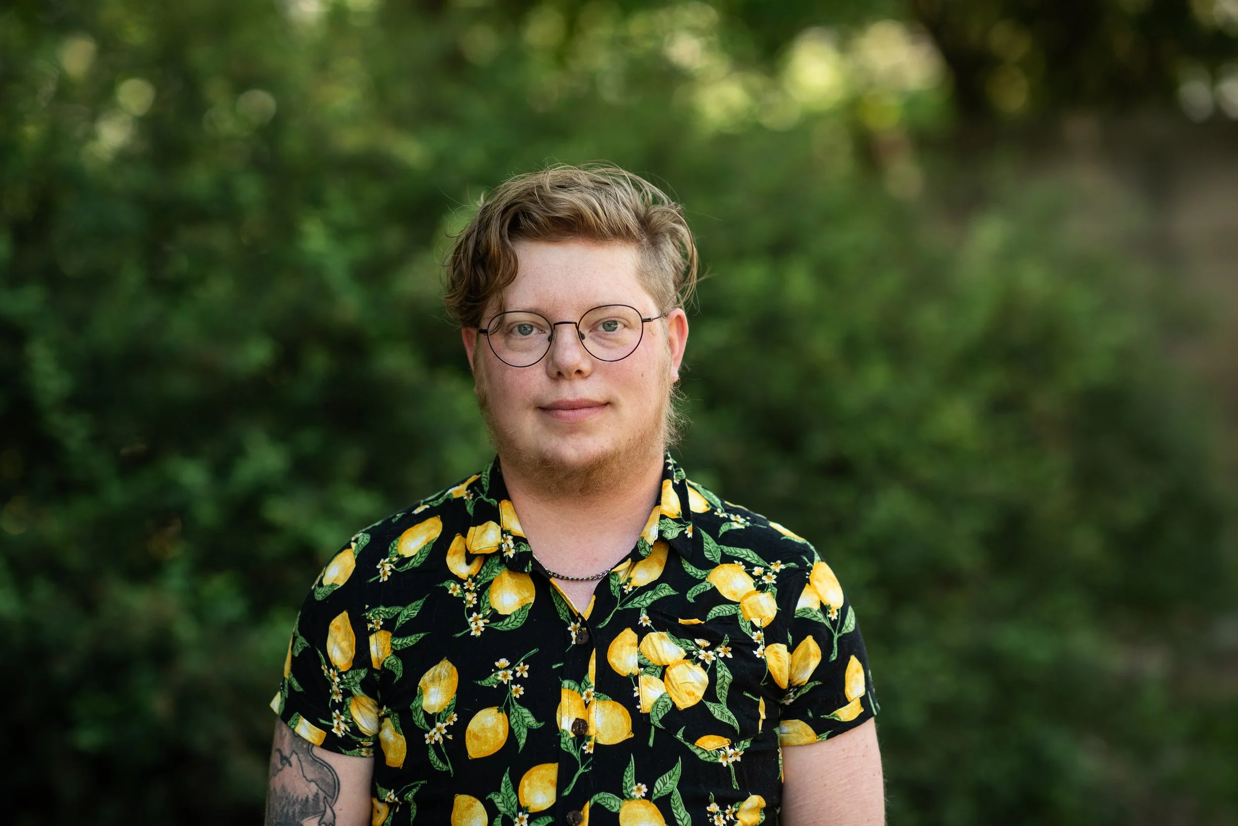 A person with short blond hair, black glasses, and weating a black shirt with a lemon design stands in front of a blurry green background.