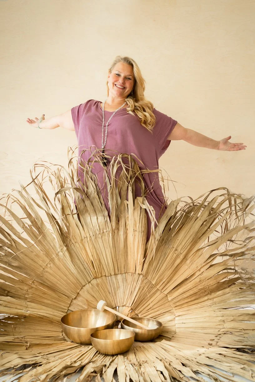 Ihminen iloisesti hymyilevä ja avoimen käsivarren asento, taustalla beige seinä, edessä suuri kuivattujen kasvien ja kultaiset kulhot.