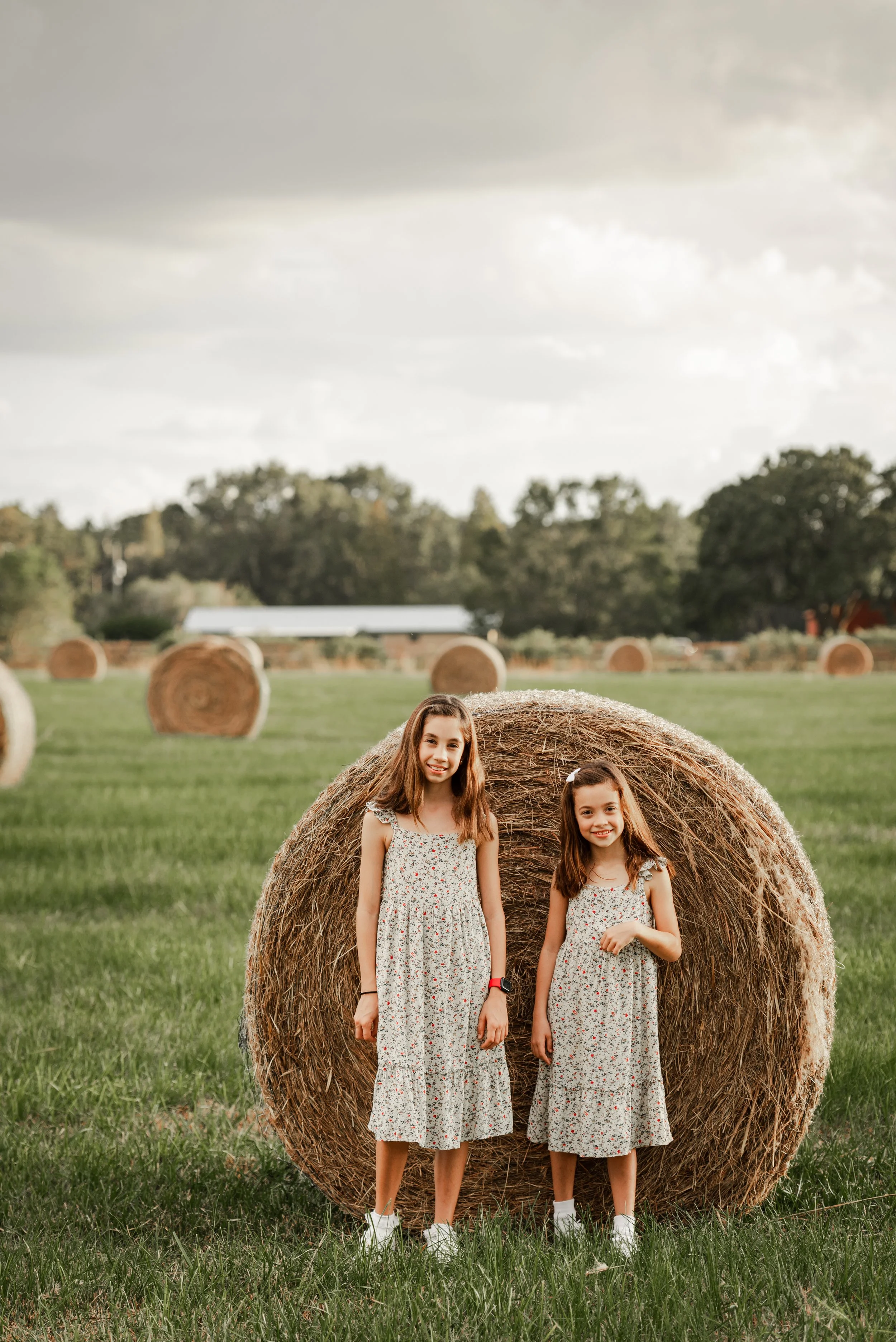 Two sisters in matching floral dresses standing in front of a large hay bale