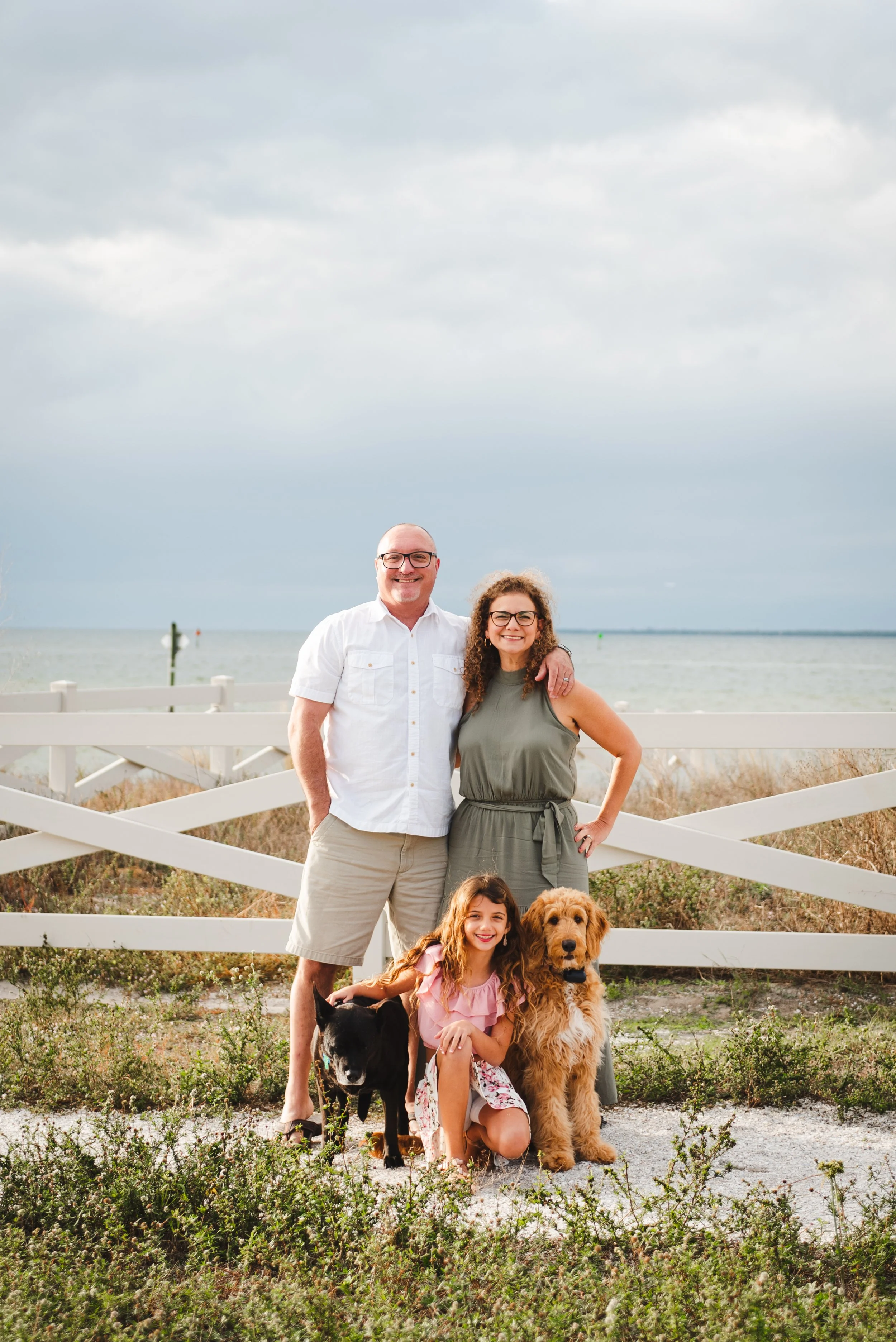 Family portrait with a senior black dog and a goldendoodle in front of a white fence at Apollo Beach Nature Preserve