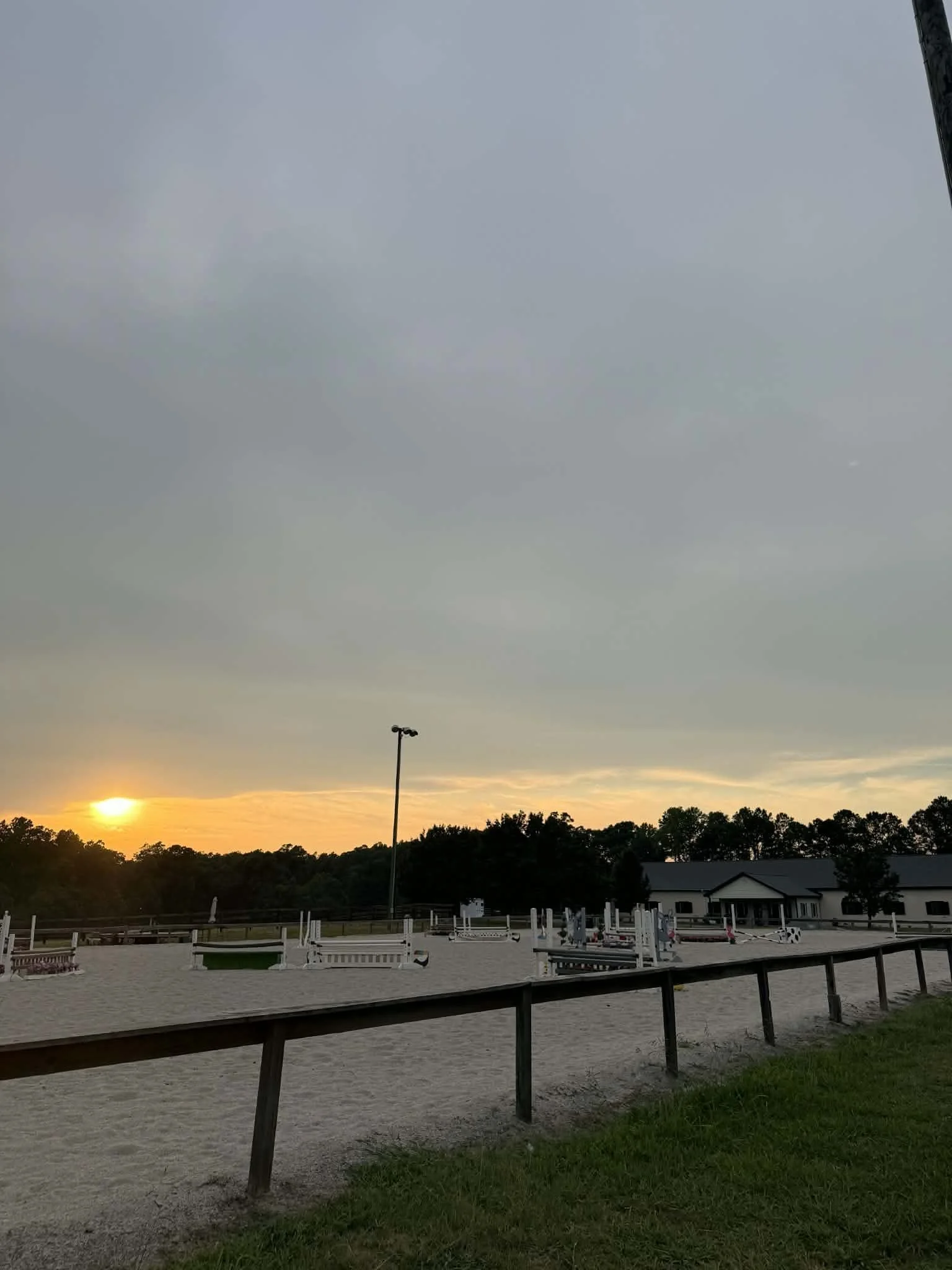 An outdoor horse riding arena at sunset with a large sandy riding area, surrounded by a wooden fence, with jumps set up for training or competition. In the background, a building and trees are visible under a cloudy sky.