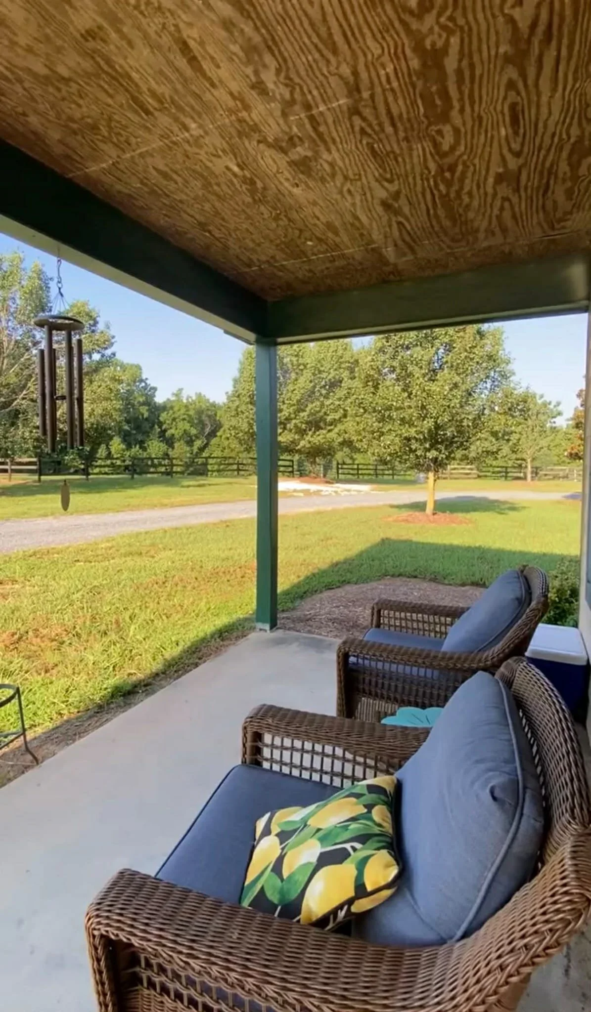 View of a covered porch with wicker chairs and cushions, overlooking a grassy yard with trees and a gravel path in the background on a sunny day.