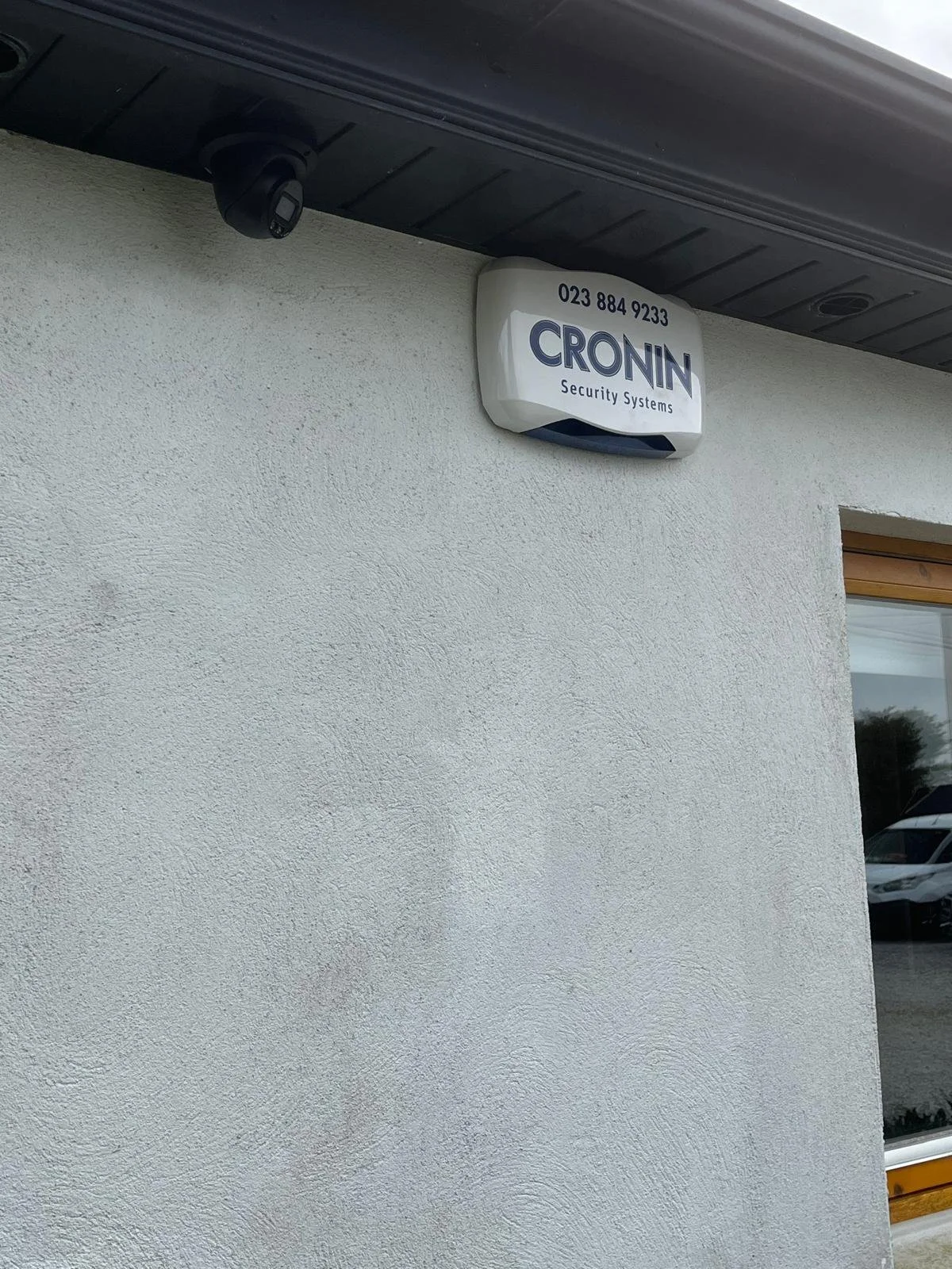 Security system sensor mounted on the exterior of a building, with a sign displaying 'CRONIN Security Systems' and a phone number.