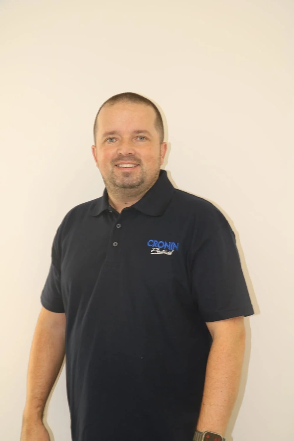 Man smiling in a black polo shirt with a company logo, standing against a plain light-colored wall.
