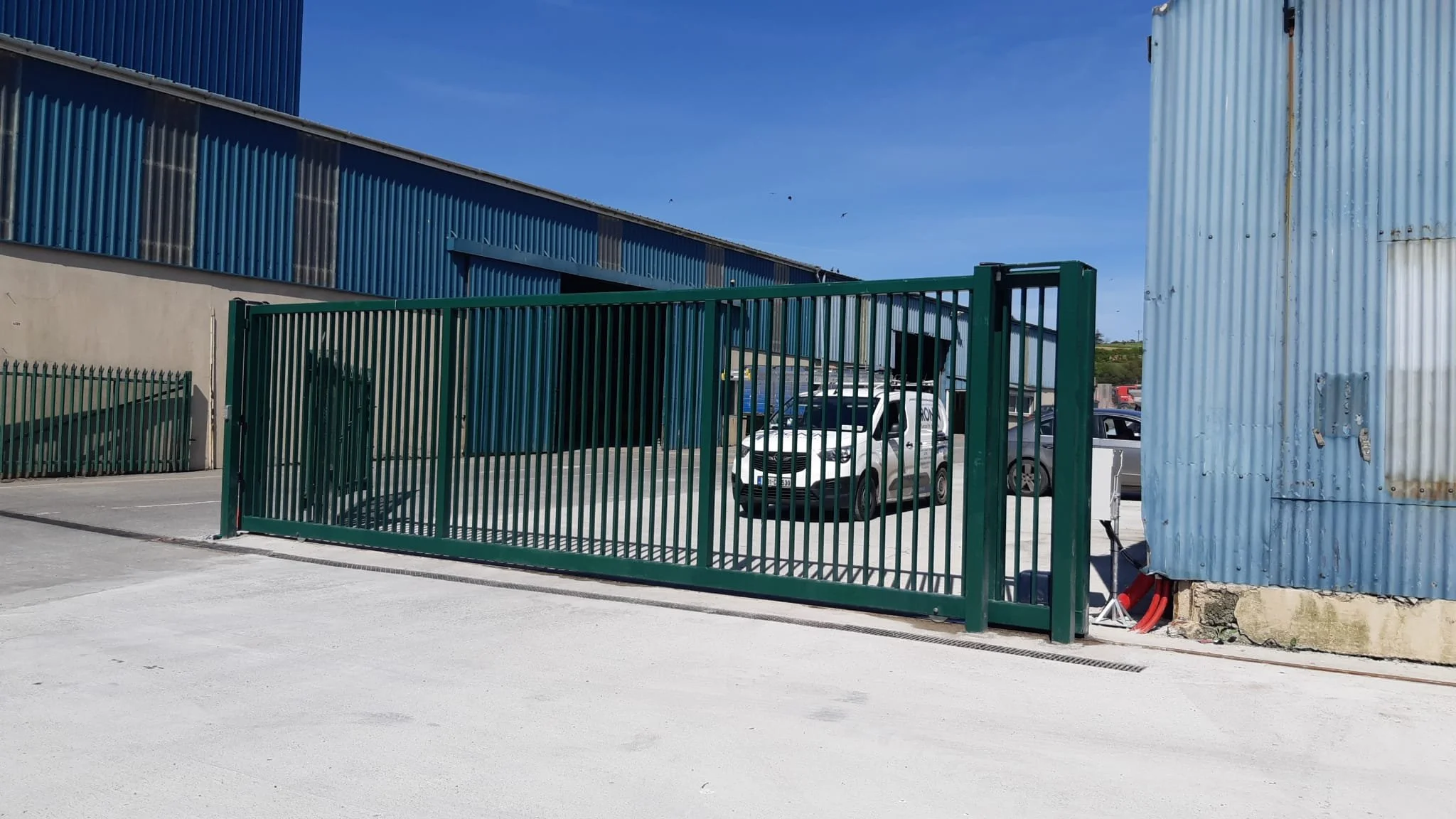 Green metal gate in front of a parking lot with several cars, surrounded by industrial buildings with blue and corrugated metal siding.
