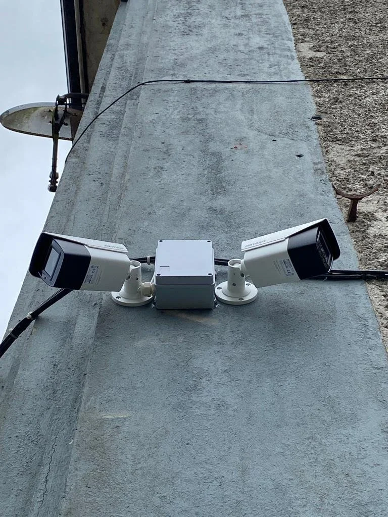 Two security cameras mounted on a concrete wall with a gray junction box between them, wires running to the cameras.