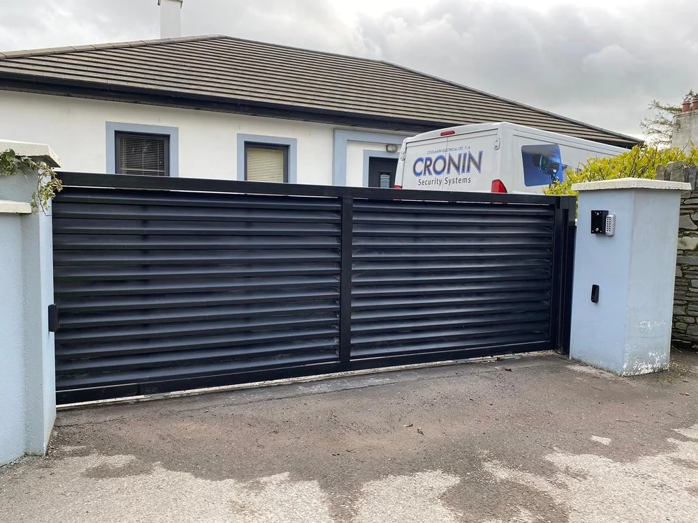 Black sliding gate opening to a driveway with a white security camera and keypad on a gray concrete pillar. A white van with the logo 'Cronin Security Systems' is parked behind the gate. A house with gray window frames and a tiled roof is in the background.