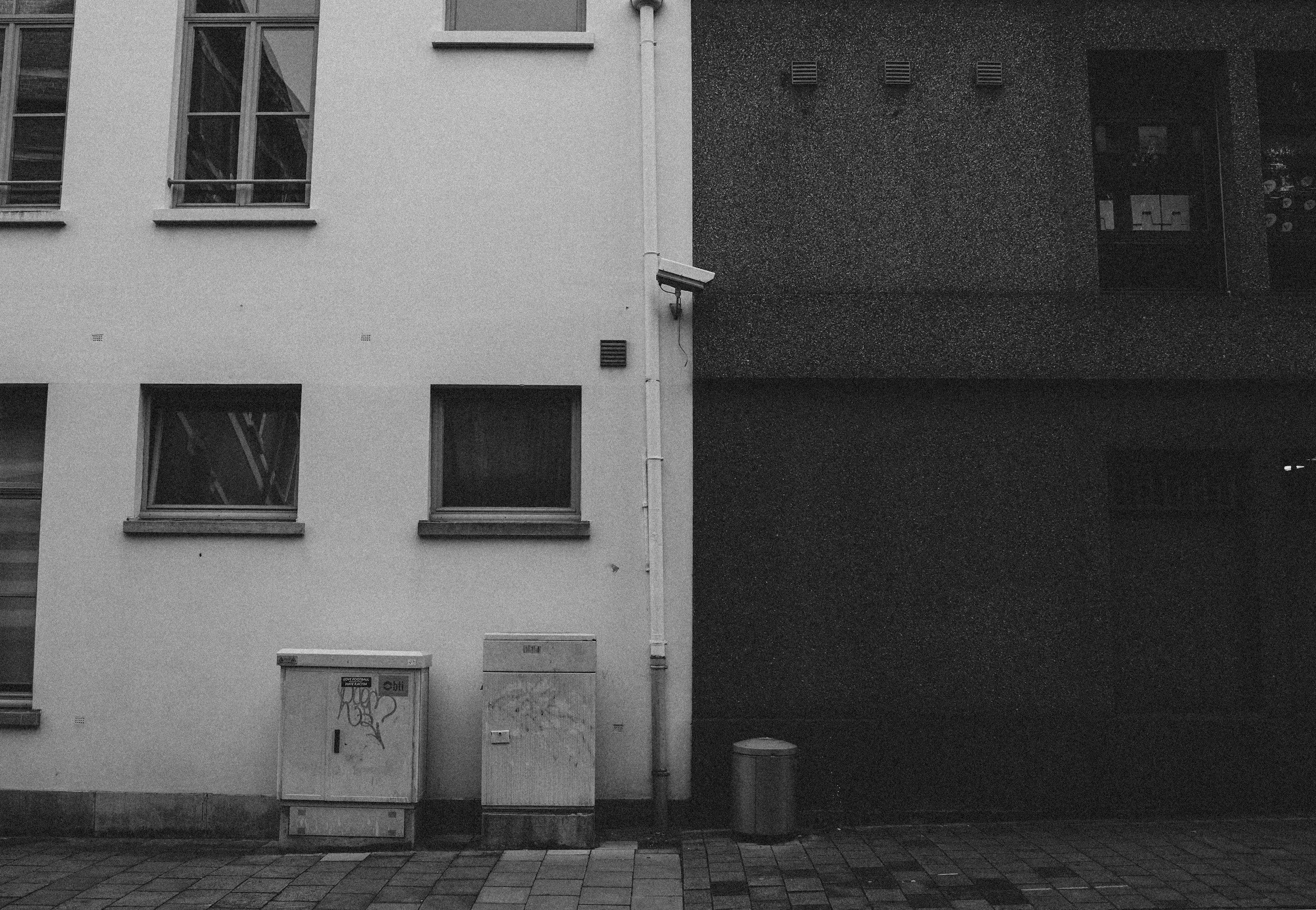 A security camera mounted under the eaves of a house with a blue tiled roof, overlooking a backyard with a wooden fence and colorful mural.