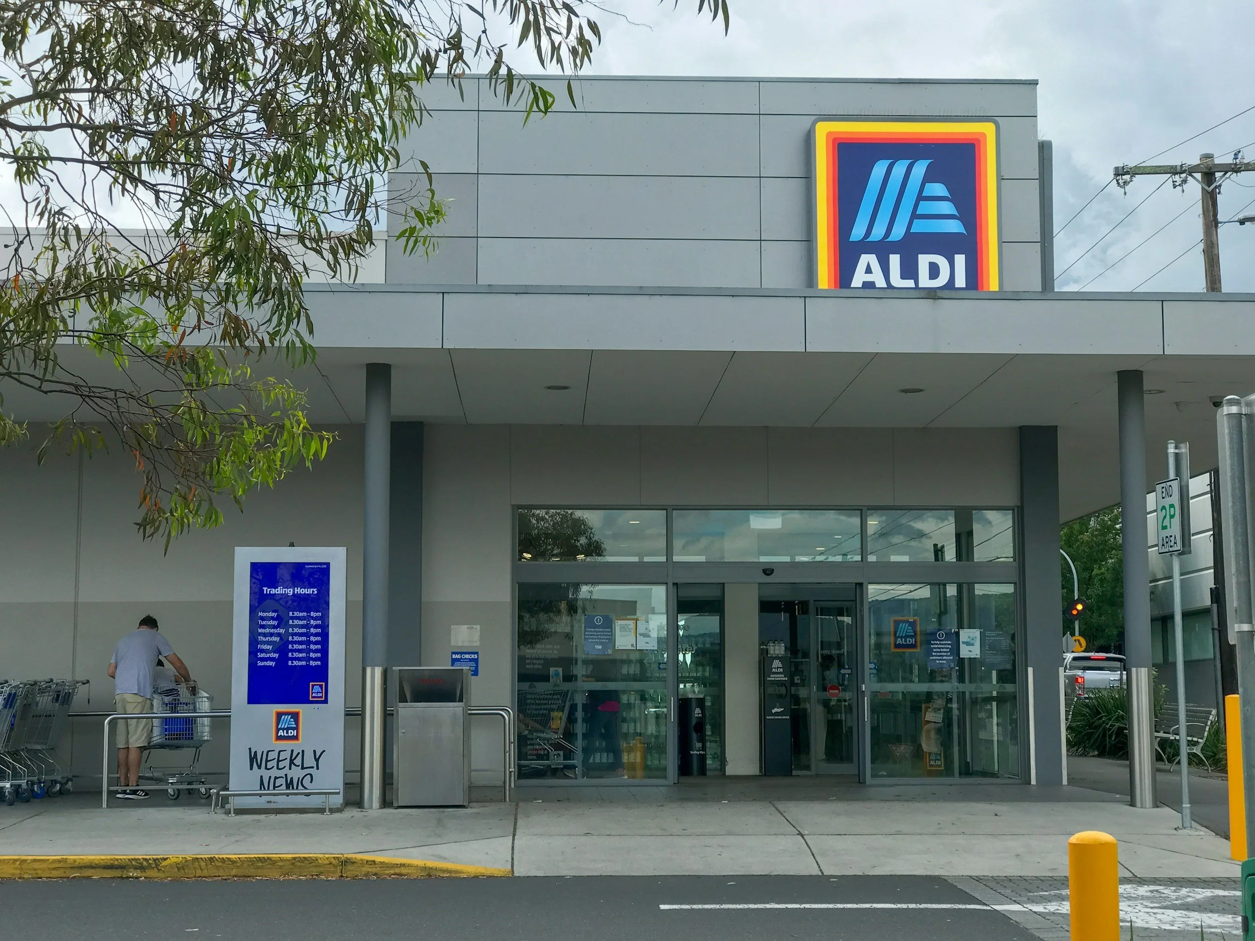 Exterior view of an ALDI grocery store entrance with shopping carts, a large trading hours sign, and a person pushing a cart near the entrance.