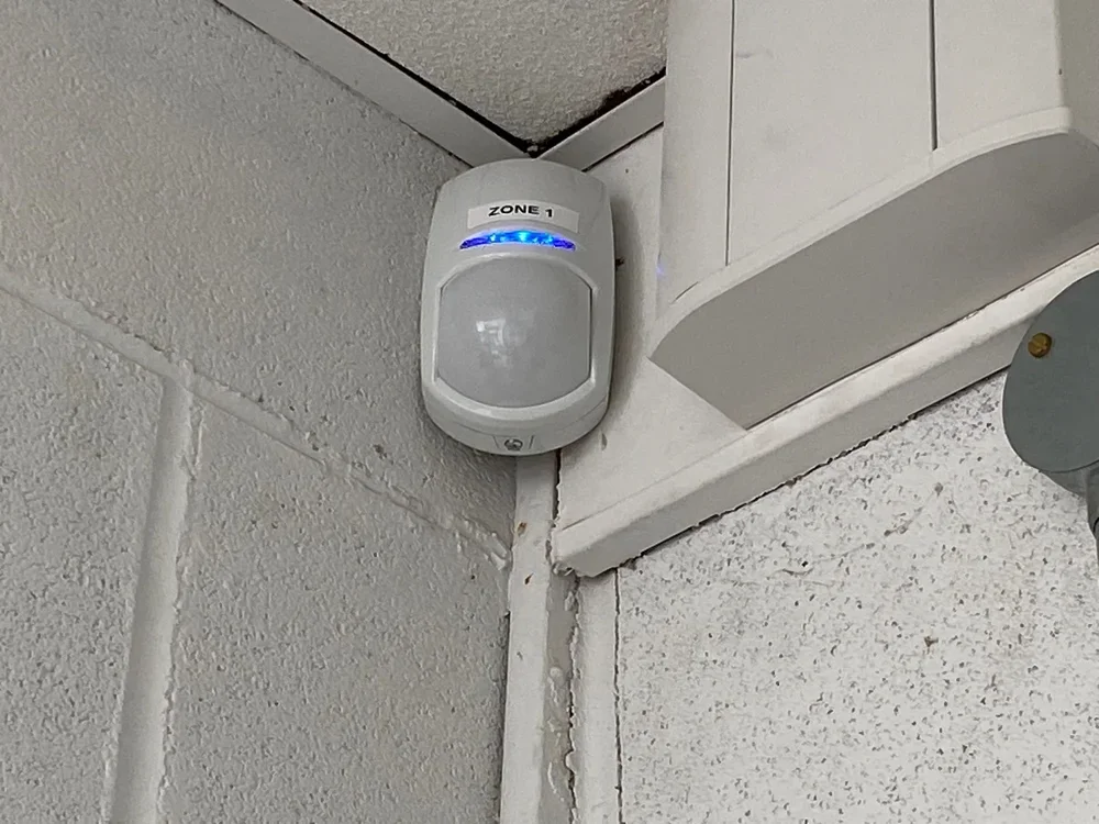 A motion detector with a blue light labeled 'ZONE 1' mounted in the corner of a room on a textured ceiling and wall.